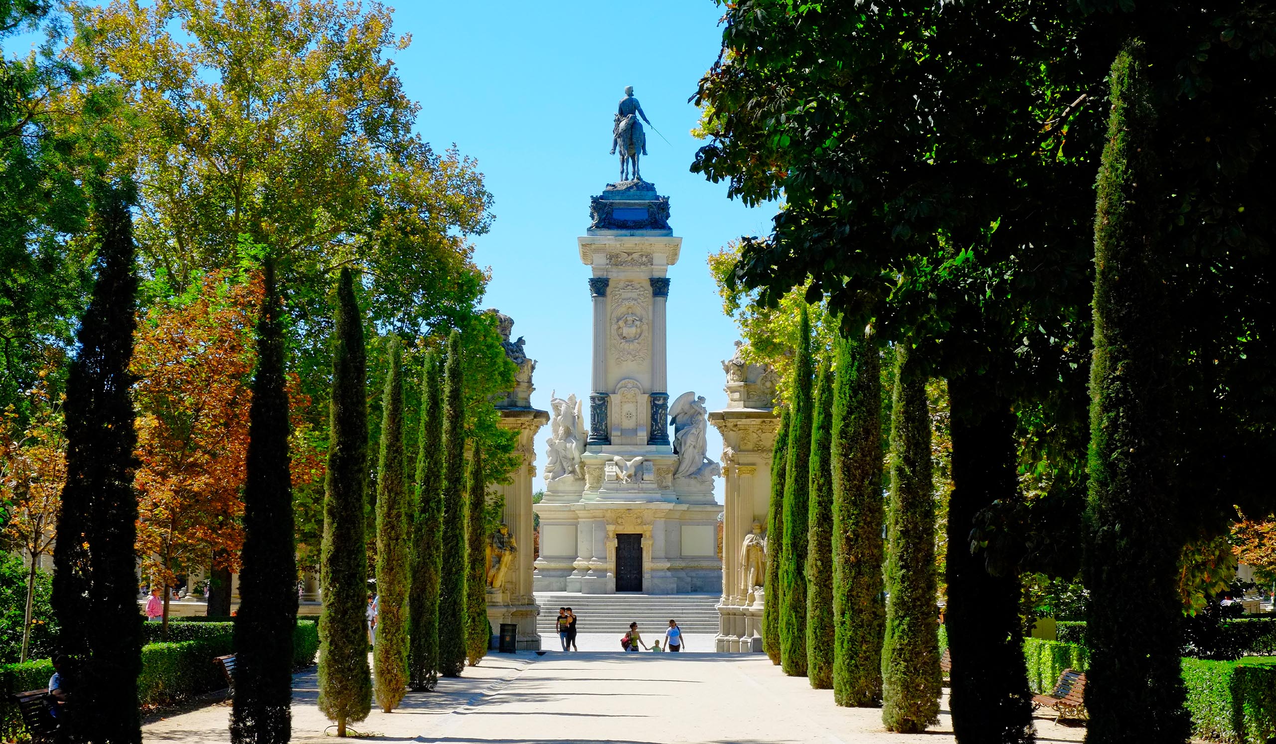 Tree-lined walkway leads to a tall monument topped with a statue, with a few people gathered at its base under a clear blue sky.