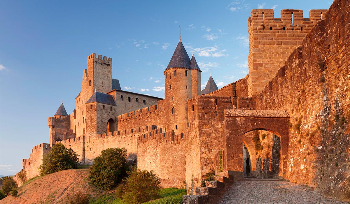 Stone fortress with towers and high walls glows in warm light, with an arched entrance along a cobblestone path.