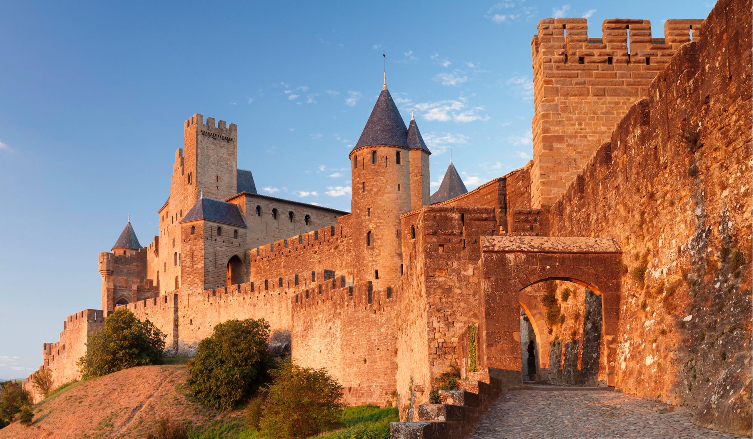 Stone fortress with towers and high walls glows in warm light, with an arched entrance along a cobblestone path.