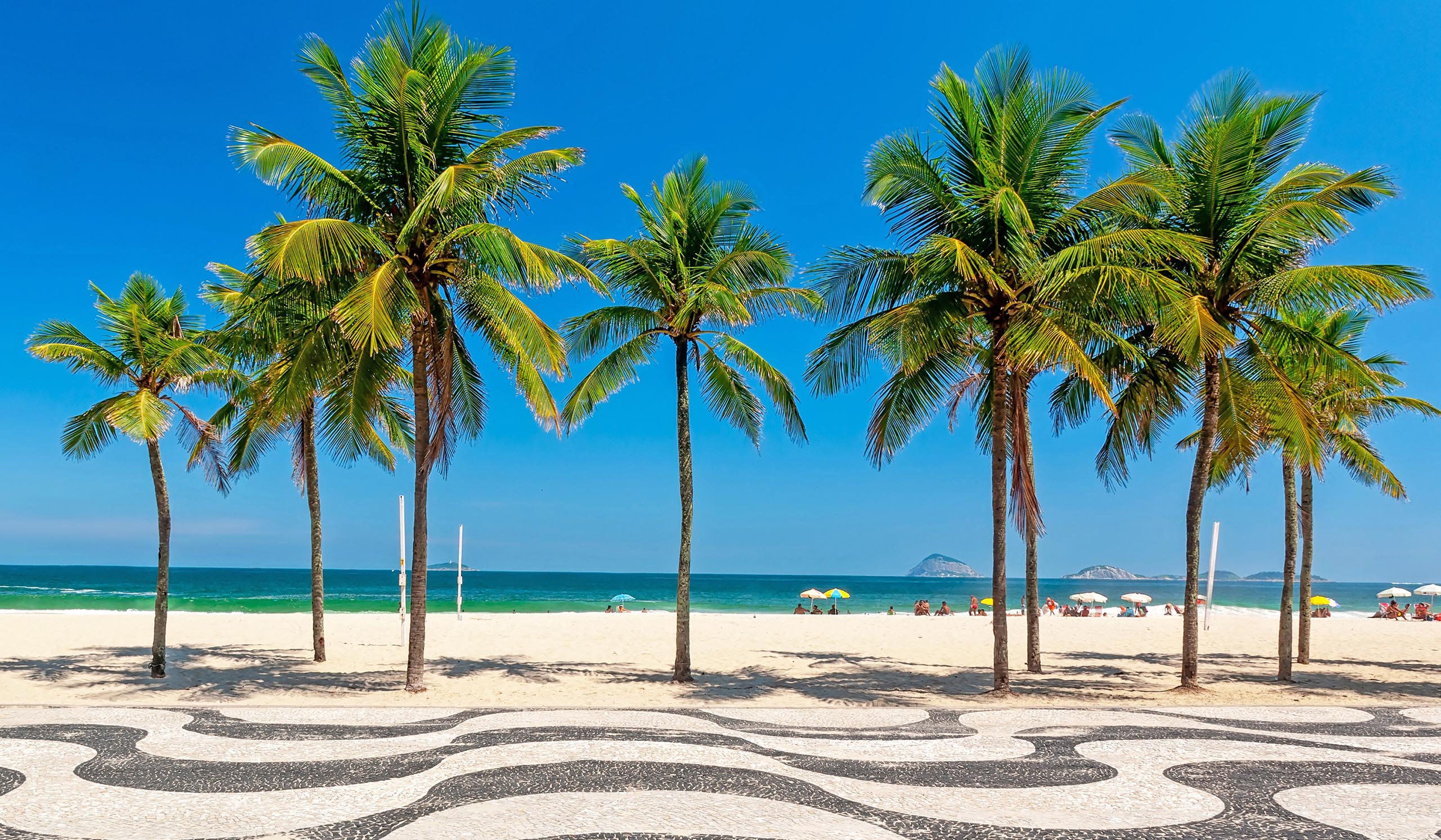 Sunny beach scene with a row of tall palm trees, overlooking white sand, turquoise water, and distant islands under a clear blue sky.
