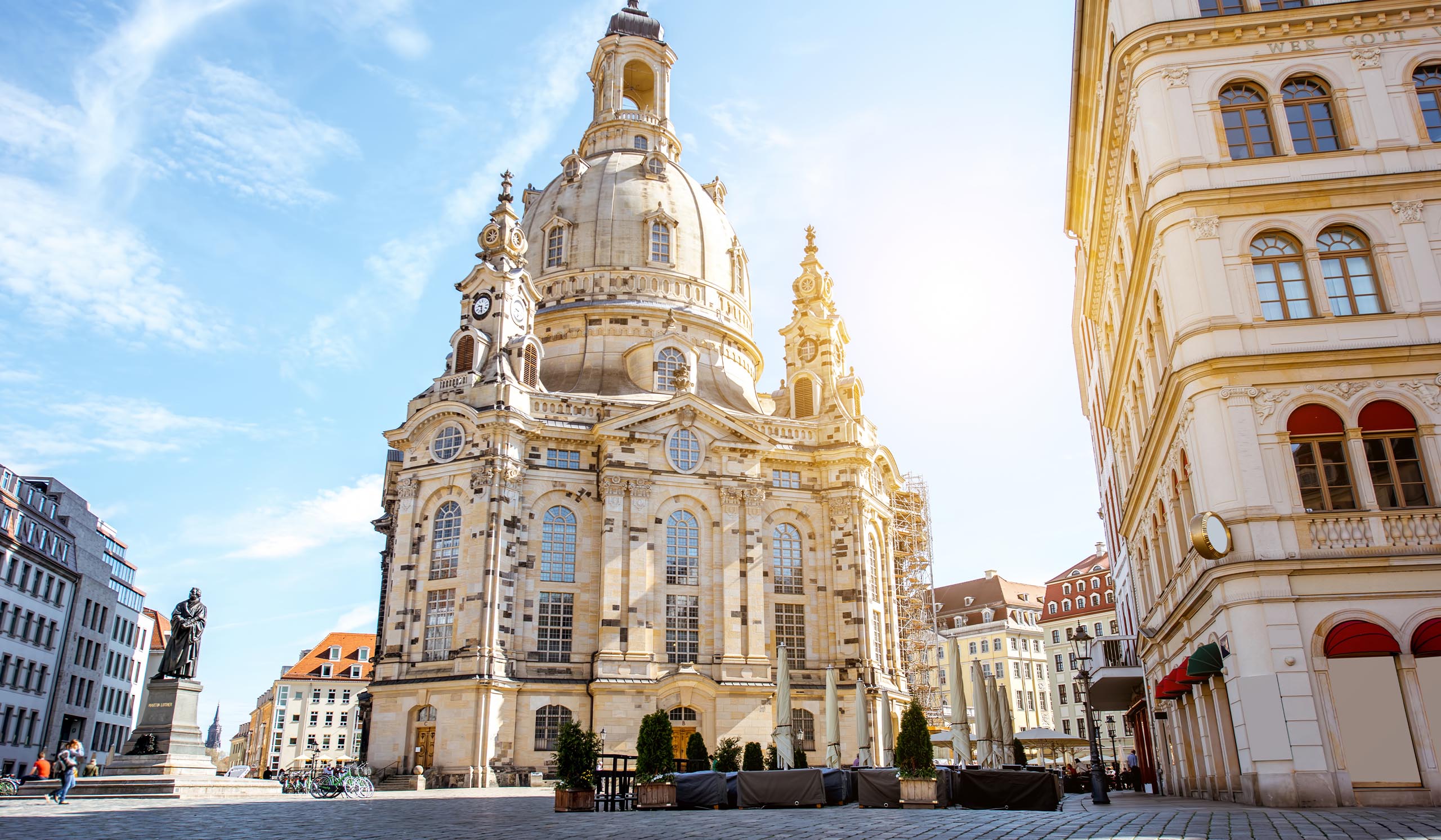 A large domed church stands in a city square, surrounded by historic buildings under a bright blue sky.
