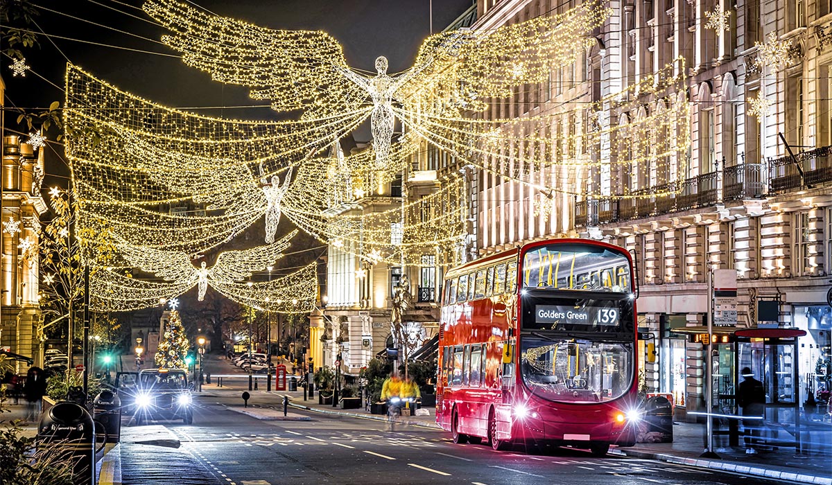 Festively lit city street at night with glowing angel-shaped holiday lights strung overhead, a red double-decker bus driving past, and illuminated buildings creating a warm, celebratory atmosphere.