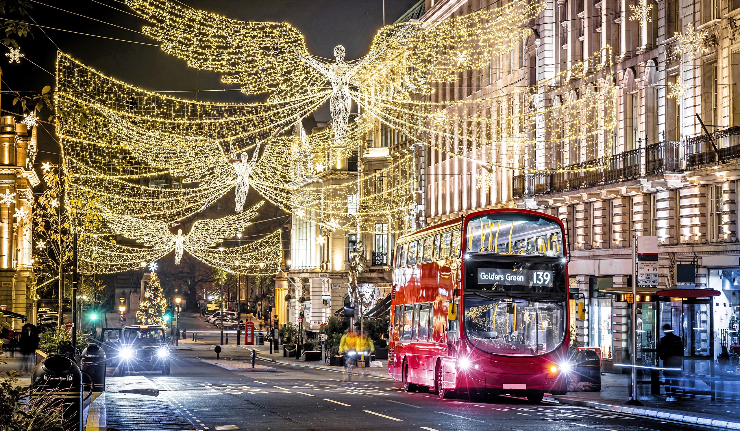 Festively lit city street at night with glowing angel-shaped holiday lights strung overhead, a red double-decker bus driving past, and illuminated buildings creating a warm, celebratory atmosphere.