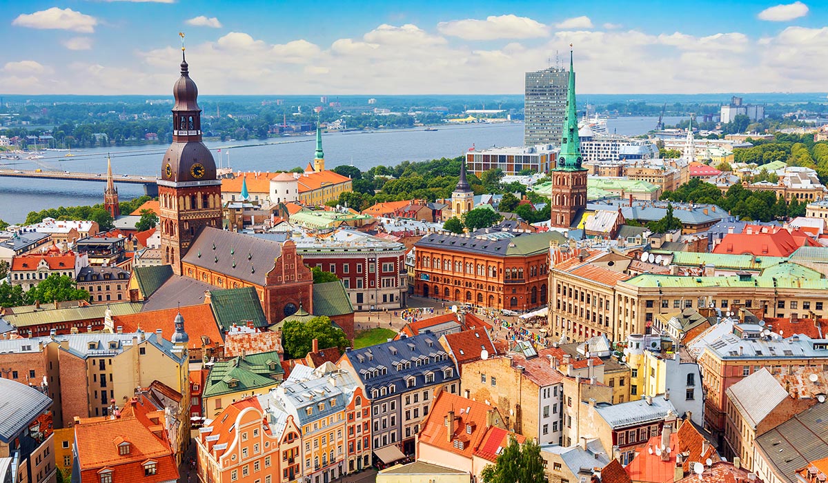 Aerial view of a colorful European city with historic buildings and church spires, including a large cathedral with a dark dome, set beside a wide river.