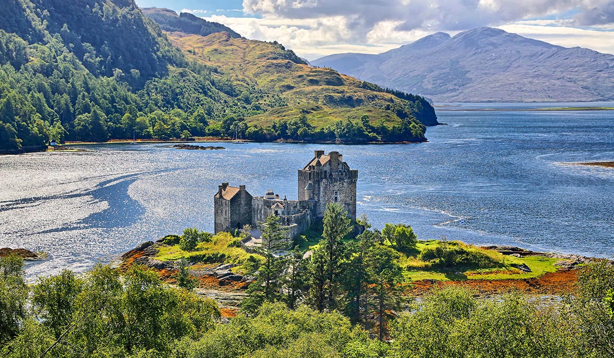 Stone castle set on a small island in a calm lake, surrounded by green hills and mountains under a partly cloudy sky.