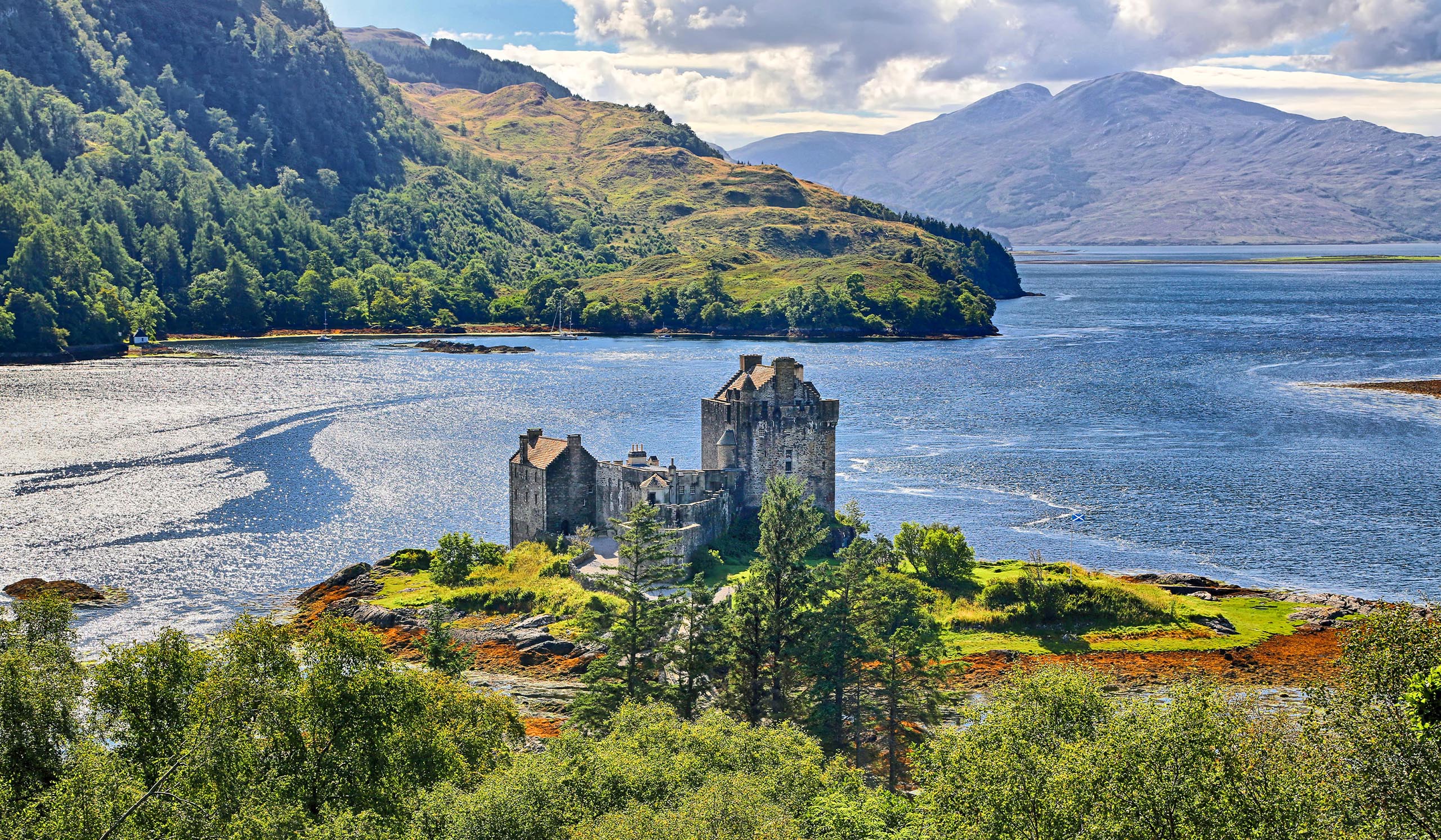 Stone castle set on a small island in a calm lake, surrounded by green hills and mountains under a partly cloudy sky.