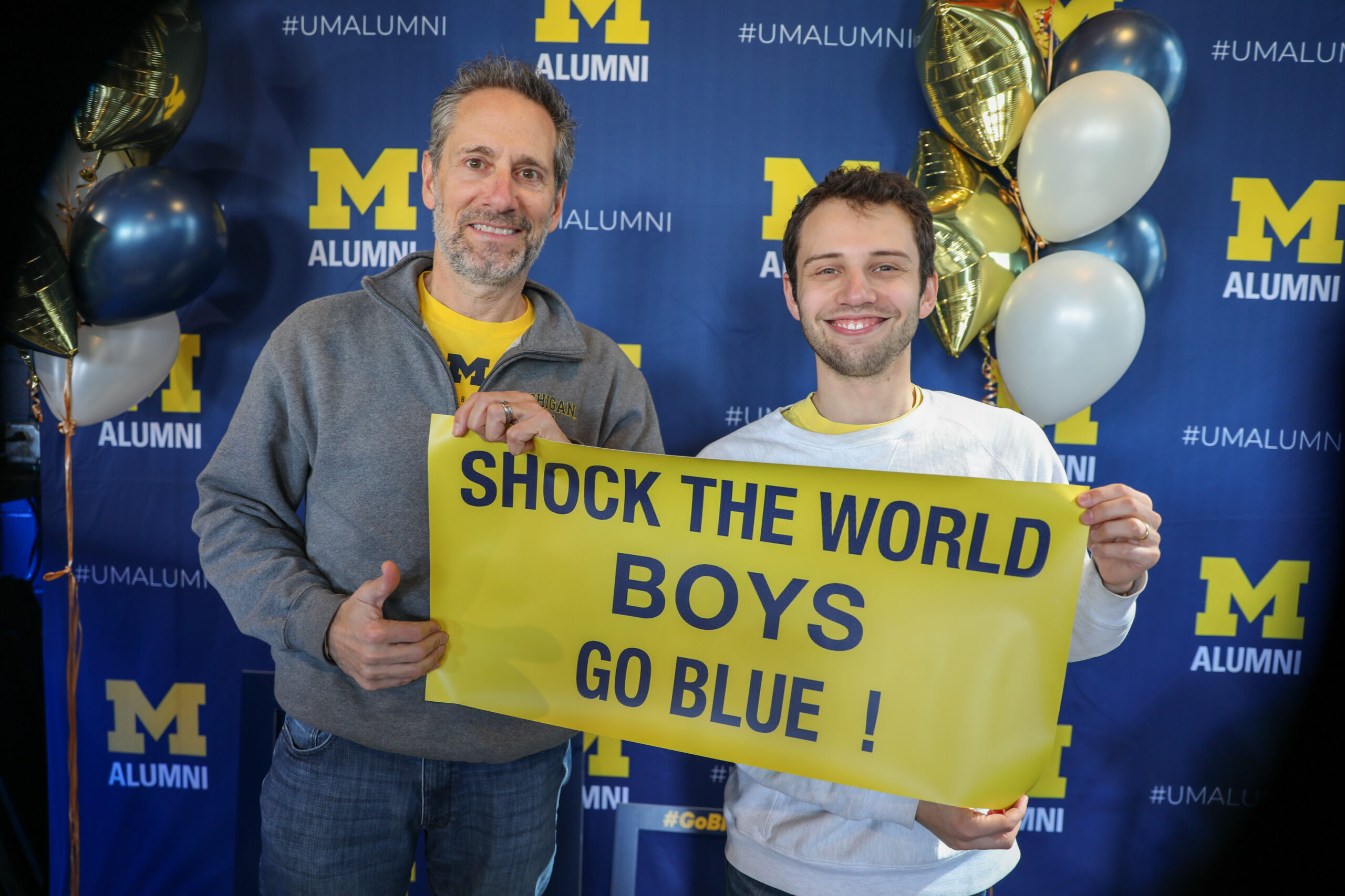 Jeffrey Gelfand (left) and his son Zac Gelfand (right) pose with the sign at the Alumni Association tailgate, wearing Michigan clothing and surrounded by Michigan decorations.