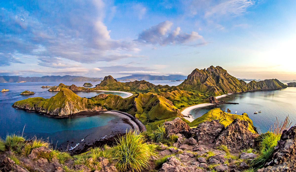 A panoramic view of lush, rugged islands with rolling green hills and curved sandy beaches surrounded by clear blue ocean, seen from an elevated rocky viewpoint.