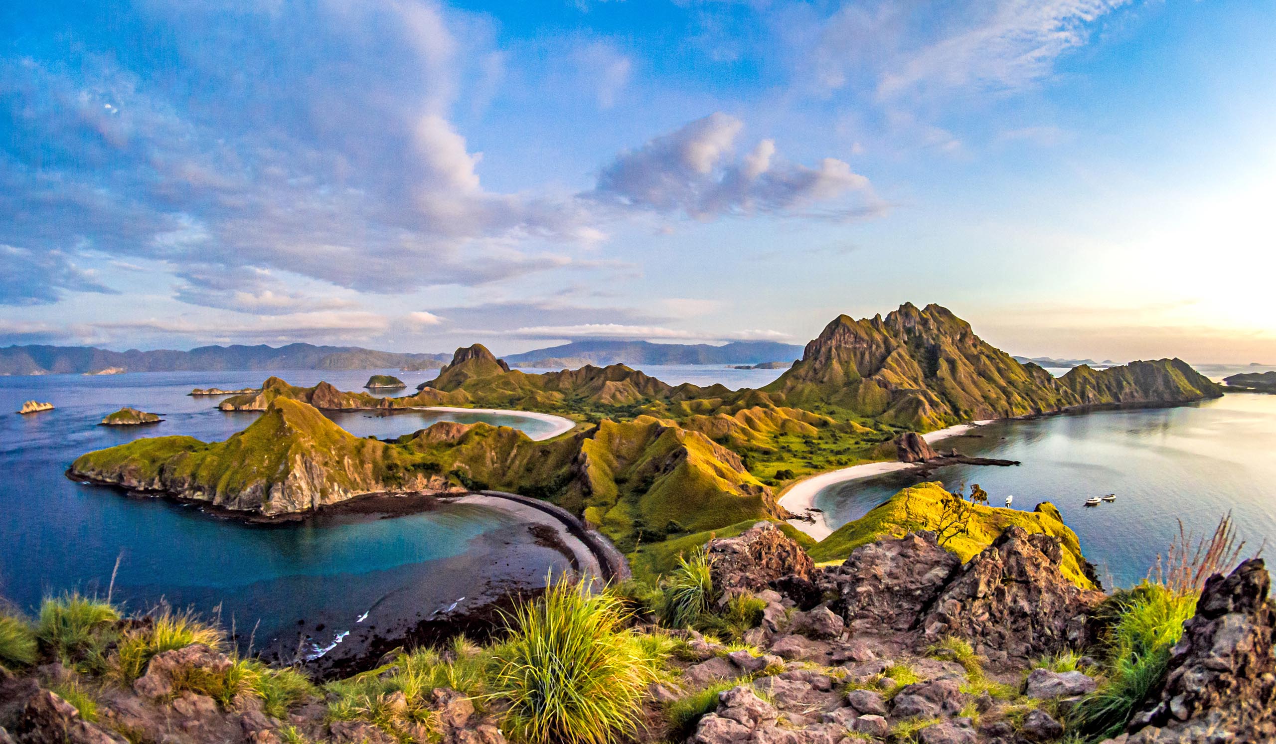 A panoramic view of lush, rugged islands with rolling green hills and curved sandy beaches surrounded by clear blue ocean, seen from an elevated rocky viewpoint.