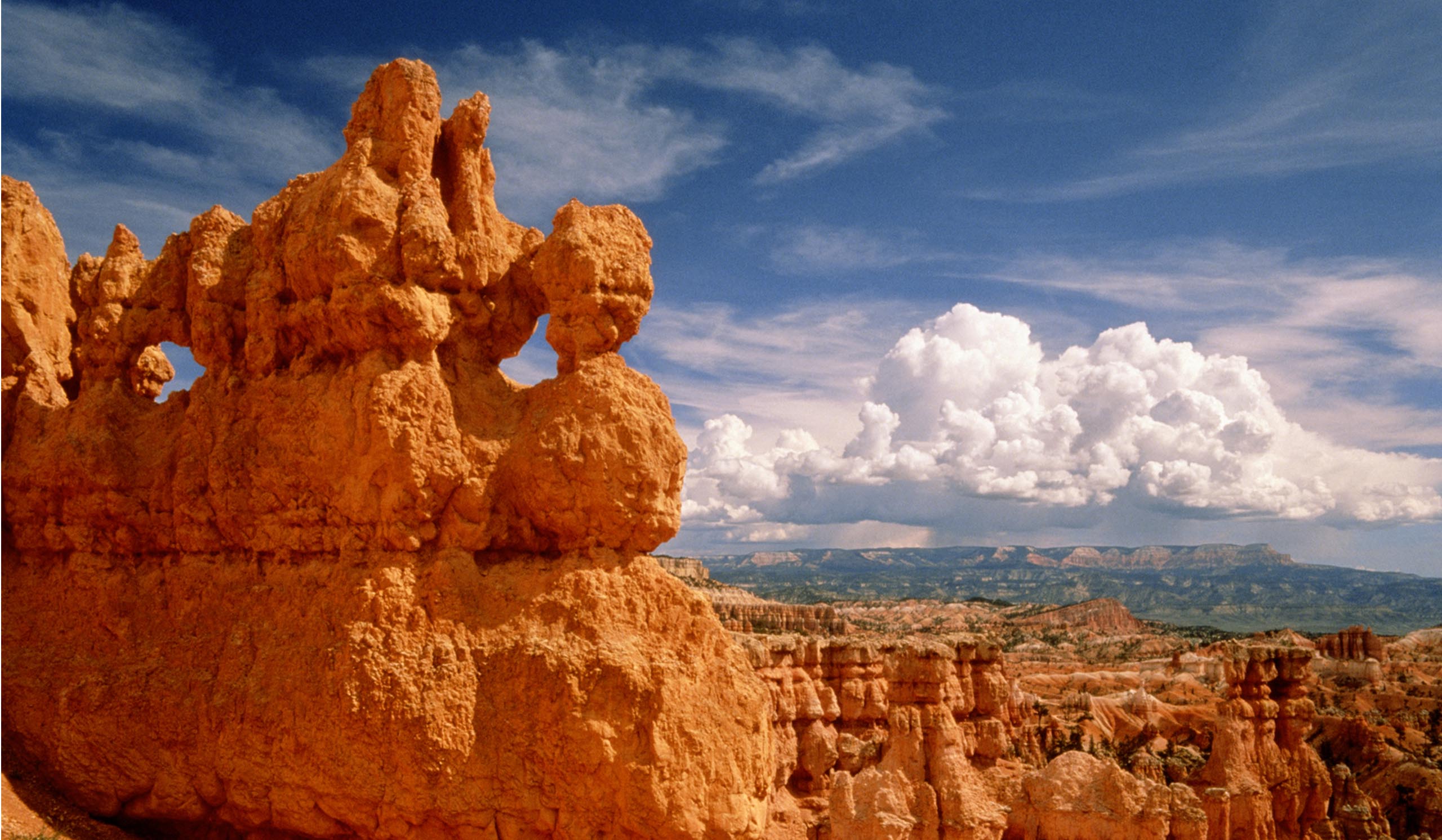 Sunlight streams over towering orange rock formations in a desert canyon, with dramatic clouds filling the sky.