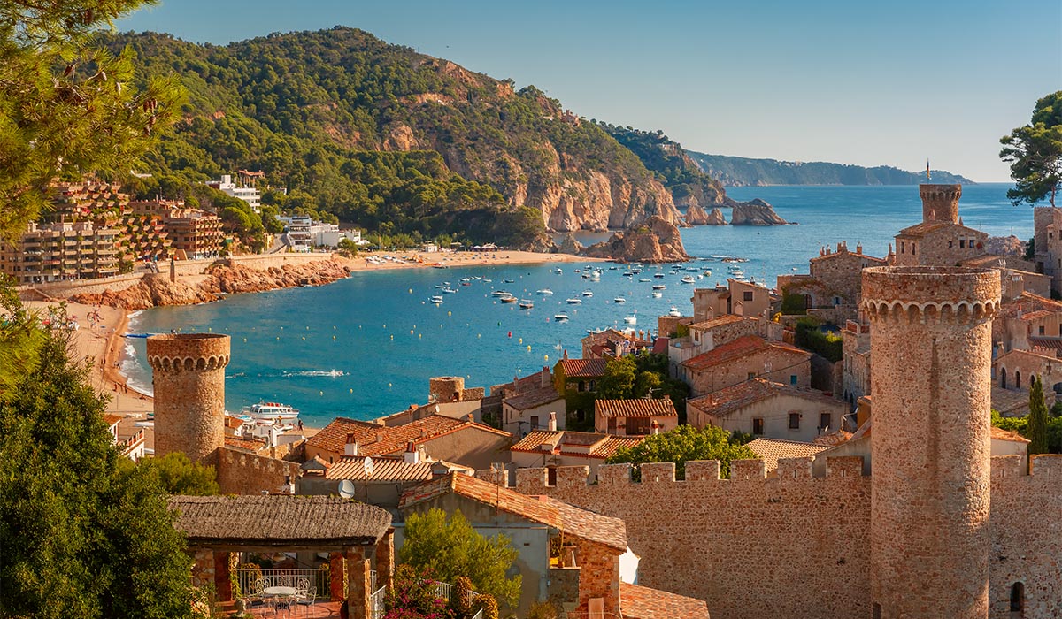 A coastal town with stone towers overlooks a curved beach and blue bay filled with small boats.