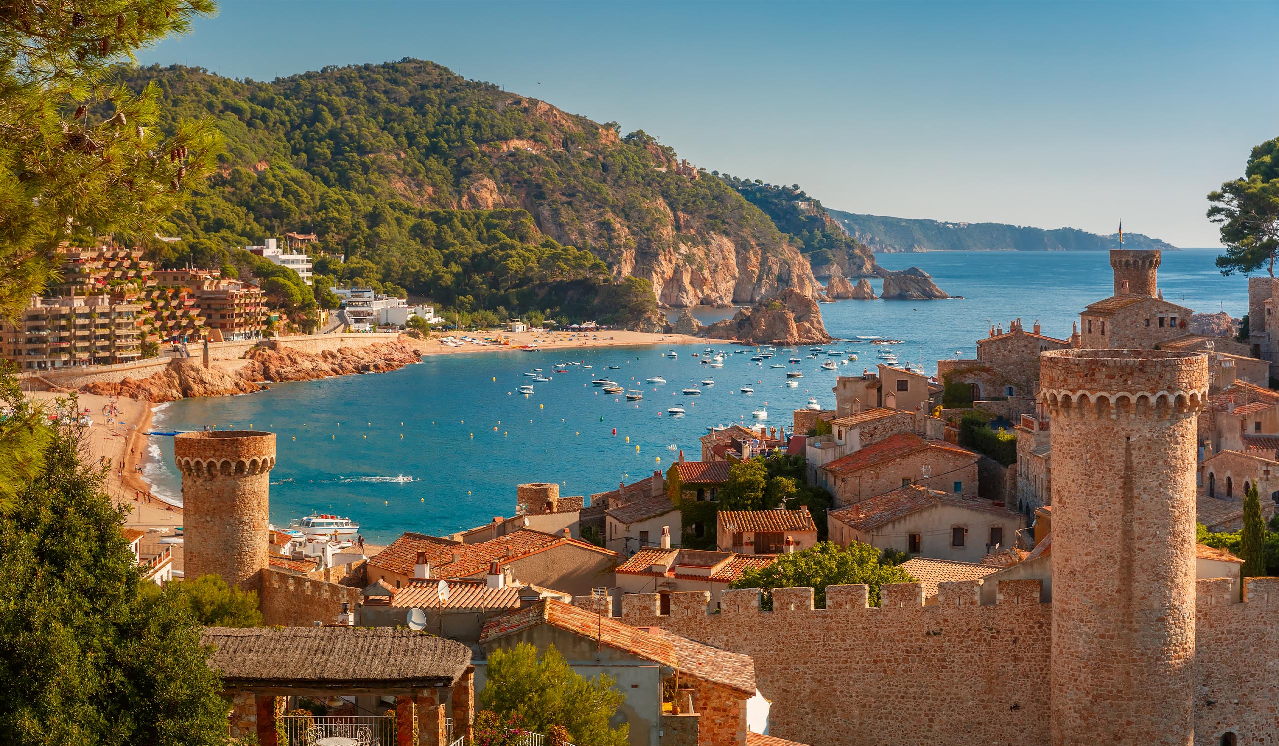 A coastal town with stone towers overlooks a curved beach and blue bay filled with small boats.