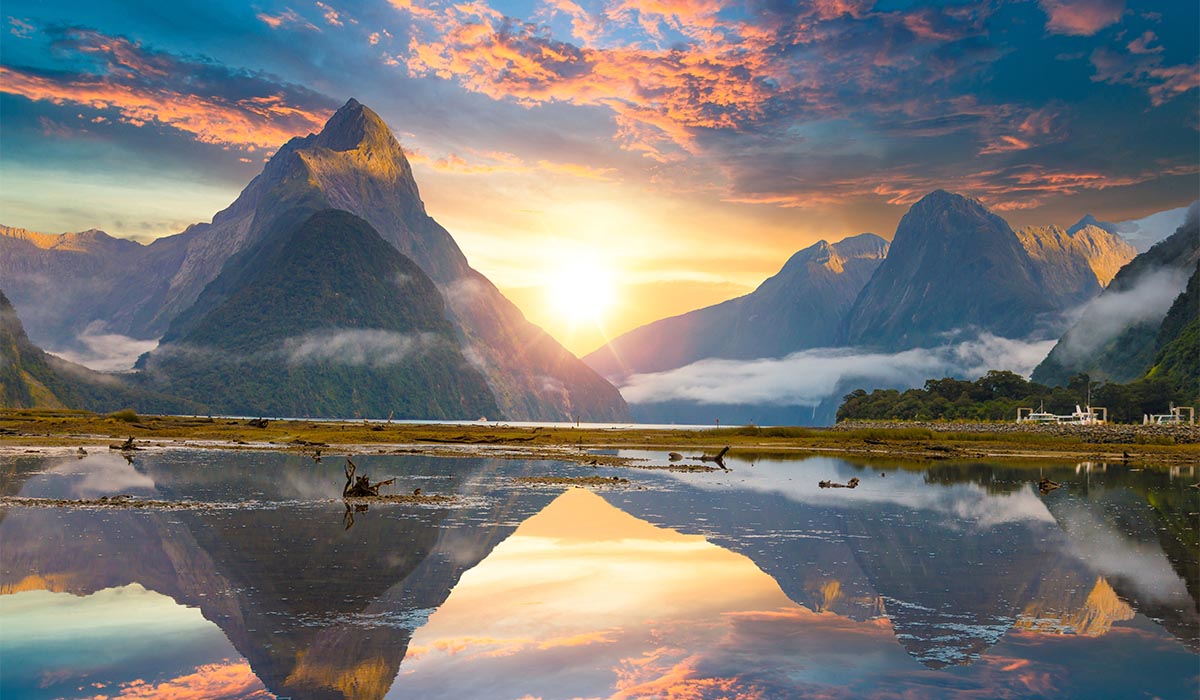 A mountain rises above a calm lake at sunrise, with colorful clouds and reflections on the water.