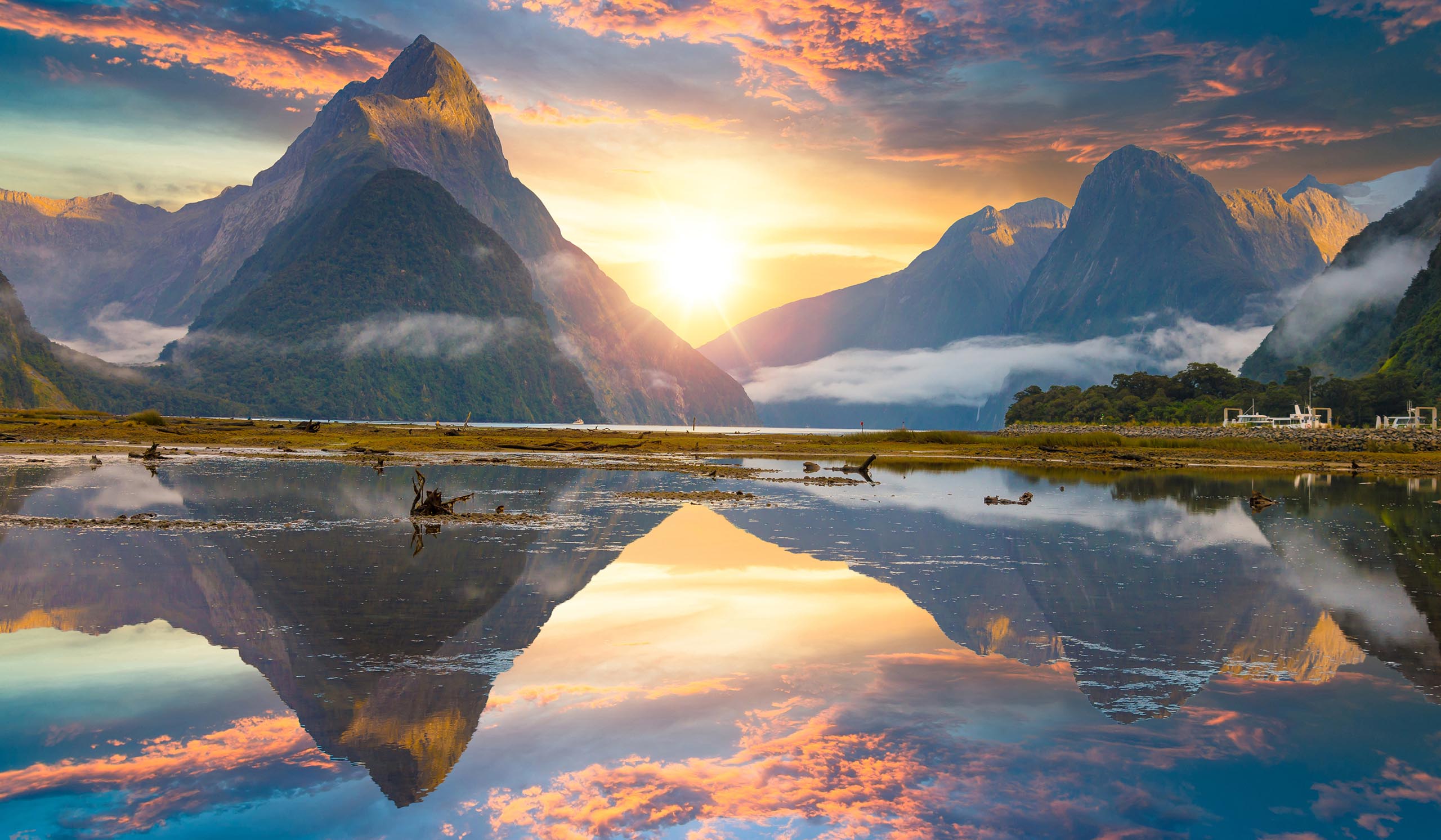 A mountain rises above a calm lake at sunrise, with colorful clouds and reflections on the water.
