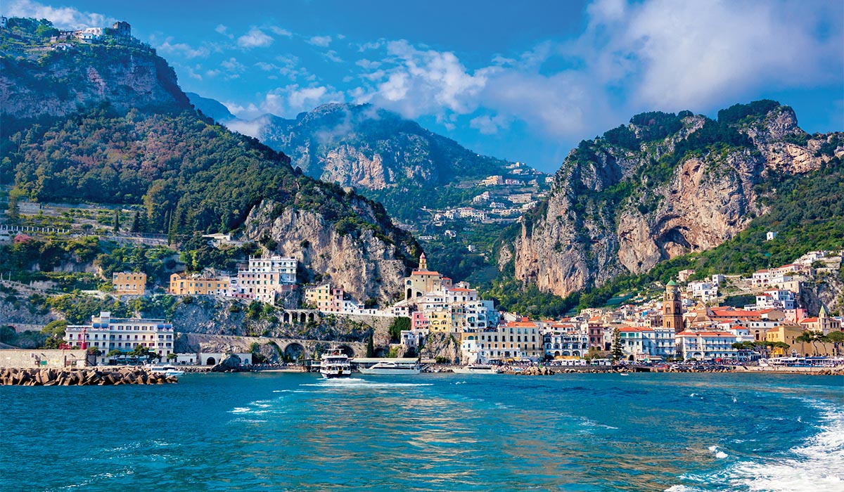 Colorful buildings line a coastal town set between steep rocky cliffs, overlooking blue water.