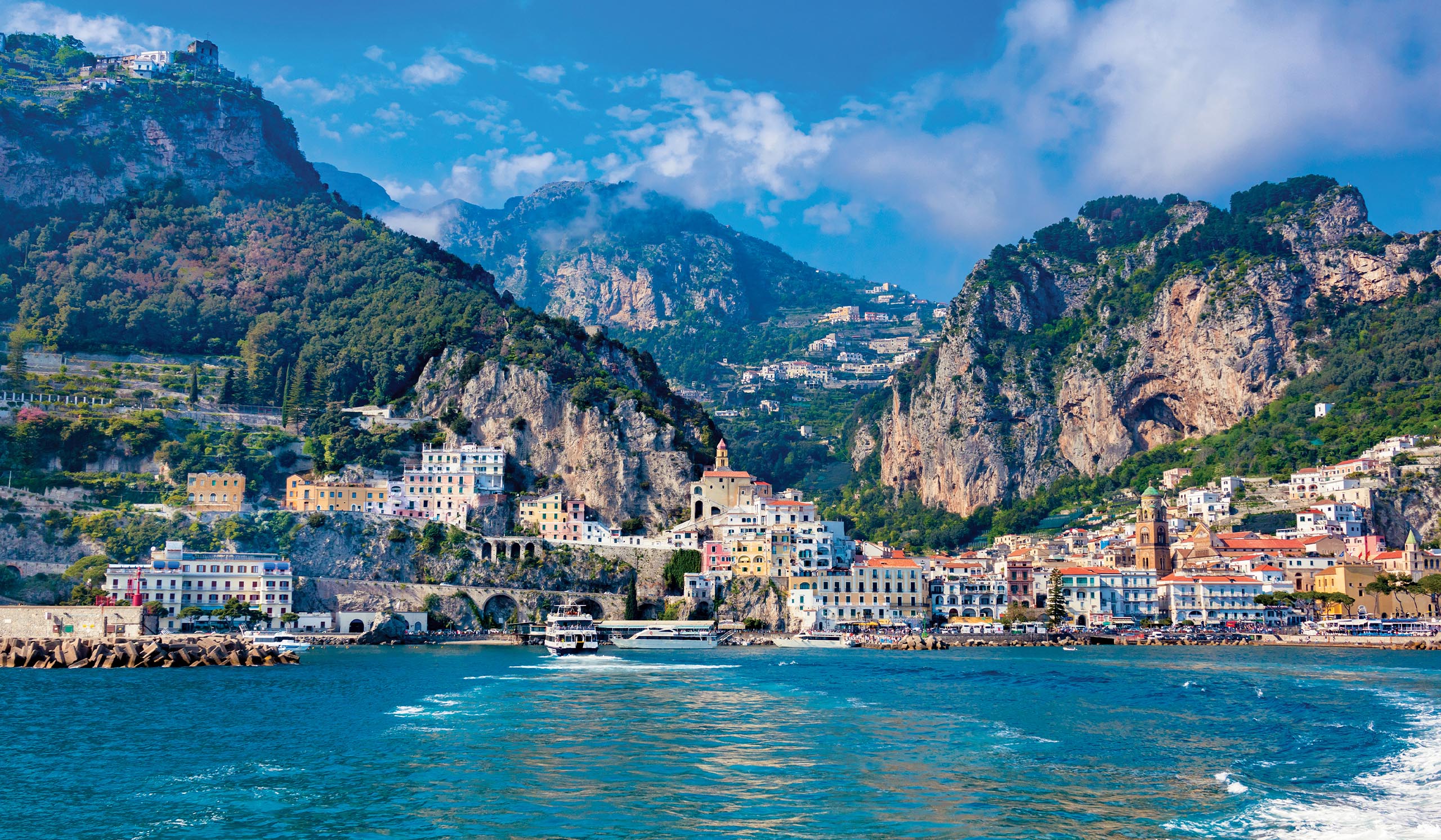 Colorful buildings line a coastal town set between steep rocky cliffs, overlooking blue water.