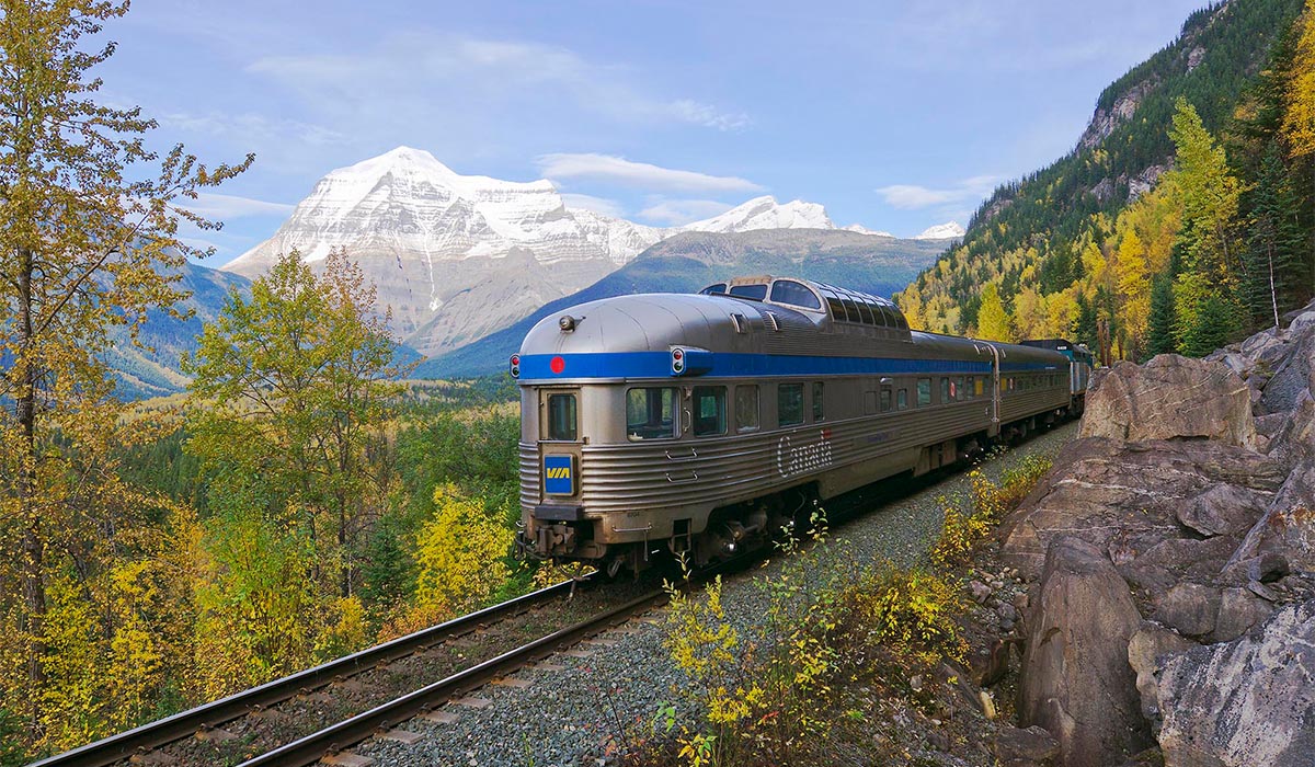 Passenger train travels along a mountainside track, with autumn trees in the foreground and snow-capped peaks rising in the distance.