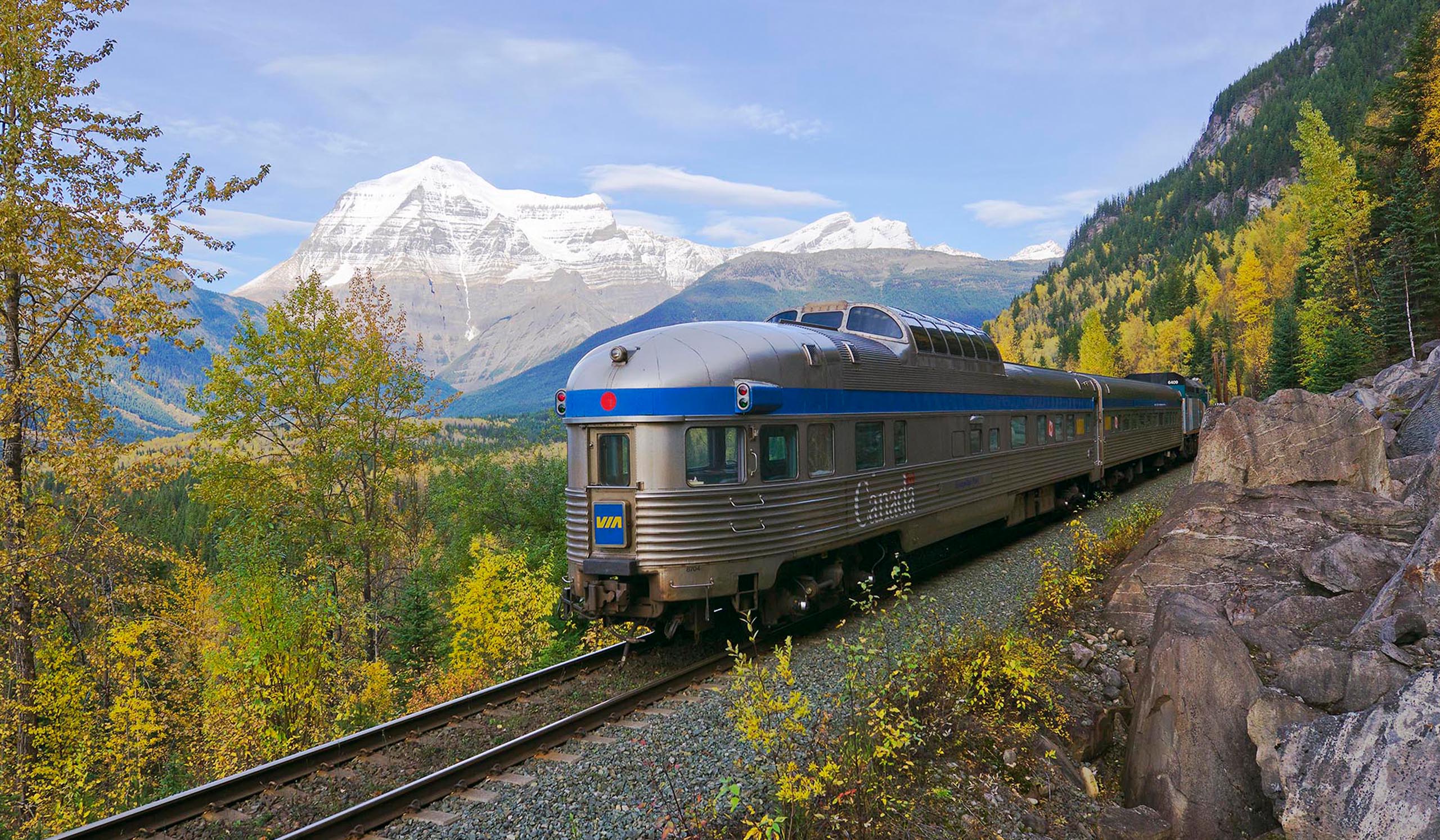Passenger train travels along a mountainside track, with autumn trees in the foreground and snow-capped peaks rising in the distance.