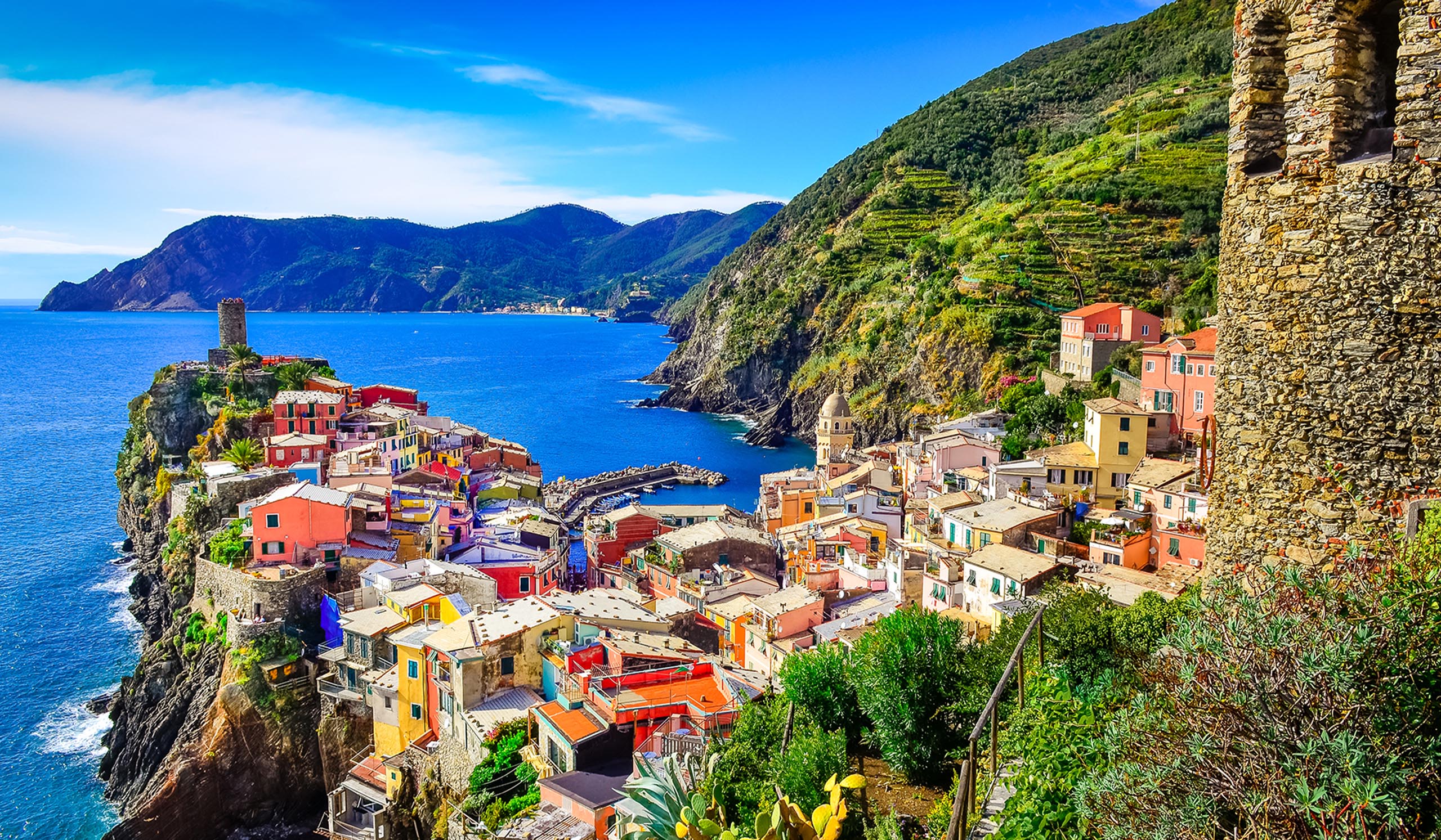 Colorful buildings cluster on a cliffside village overlooking the blue sea, with green hills rising behind.