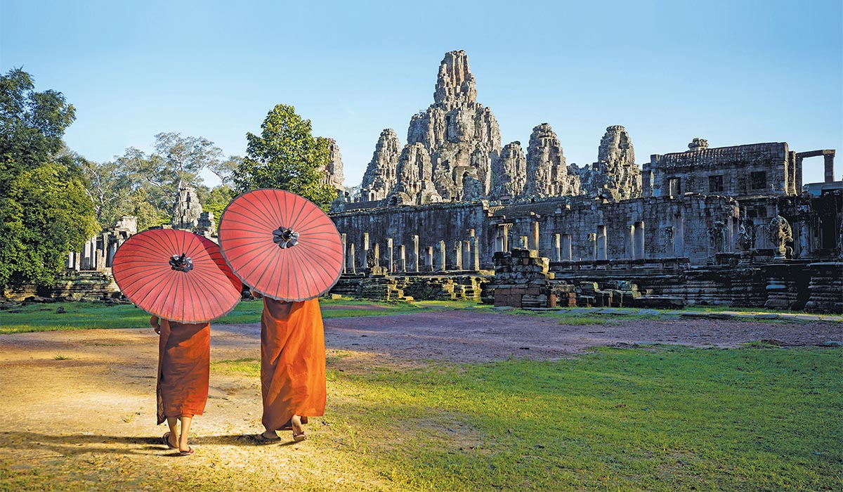 Two people in traditional robes walk with red umbrellas in front of ancient stone temple ruins.