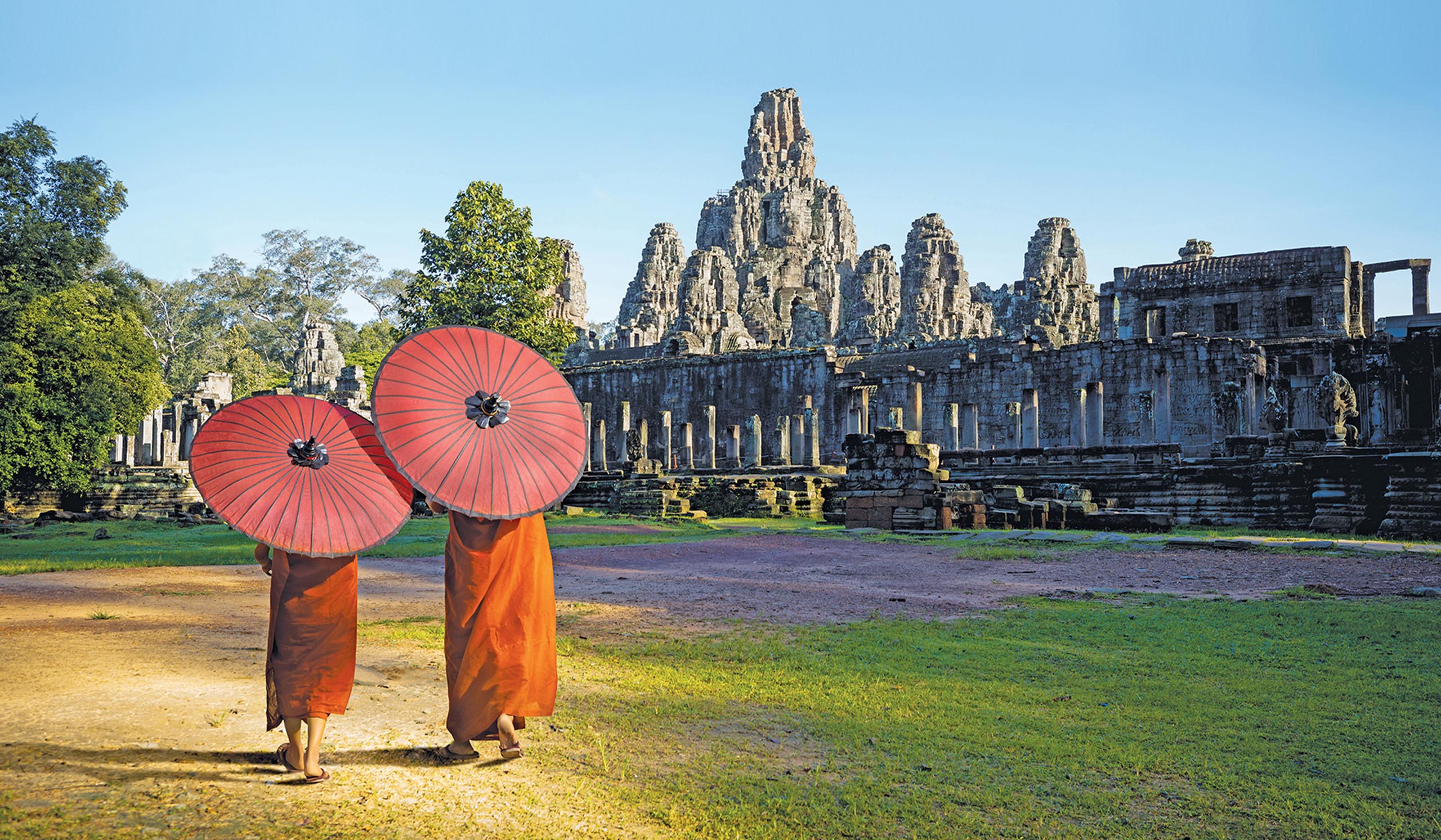 Two people in traditional robes walk with red umbrellas in front of ancient stone temple ruins.