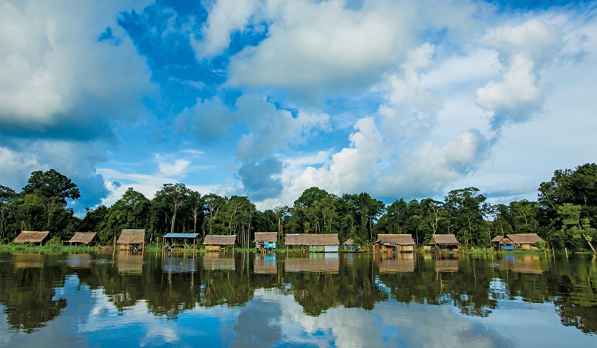 Riverside village with small thatched huts lining the water’s edge, backed by dense tropical forest and mirrored in the calm, reflective river beneath a wide, cloud-filled sky.