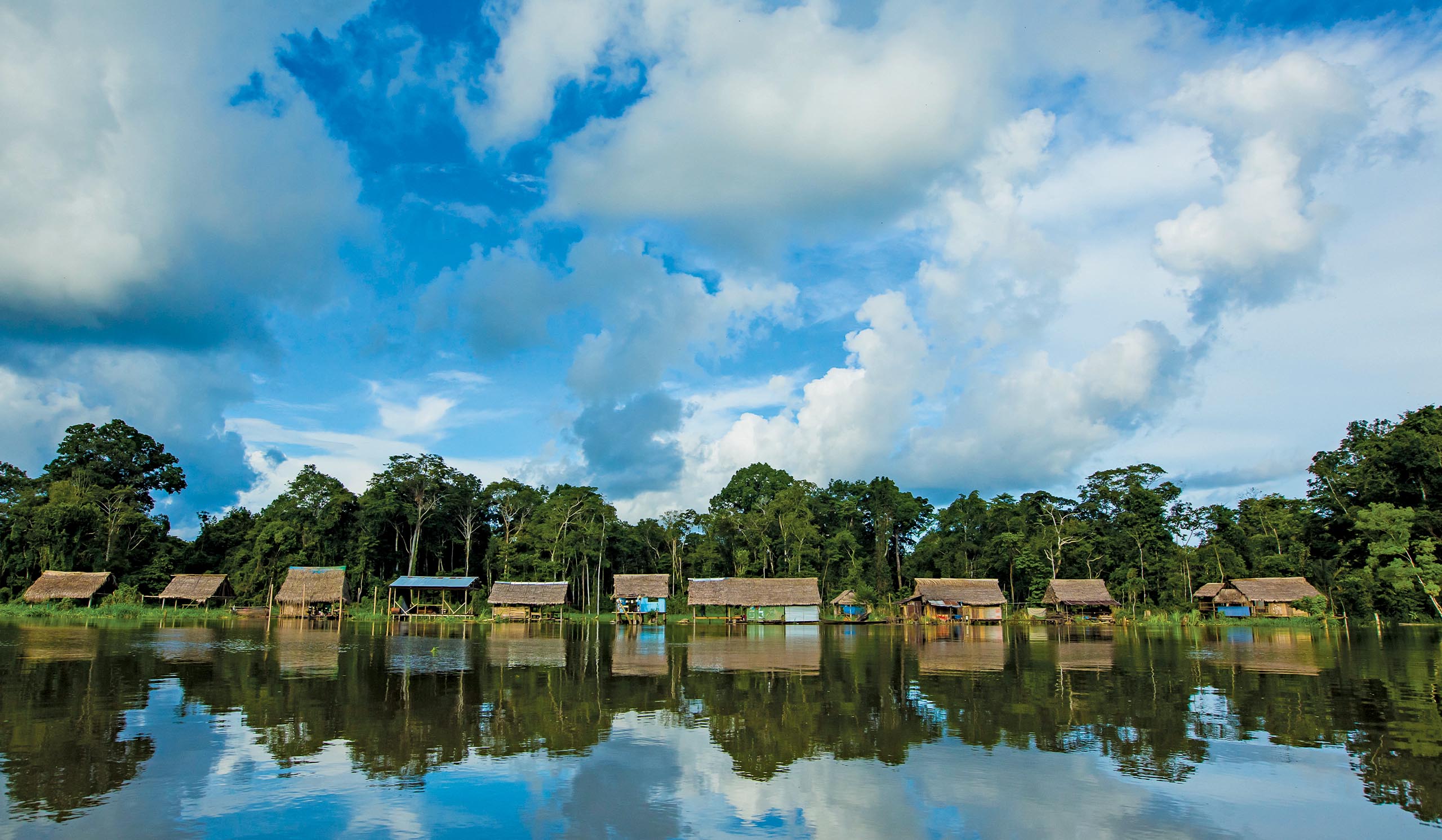 Riverside village with small thatched huts lining the water’s edge, backed by dense tropical forest and mirrored in the calm, reflective river beneath a wide, cloud-filled sky.