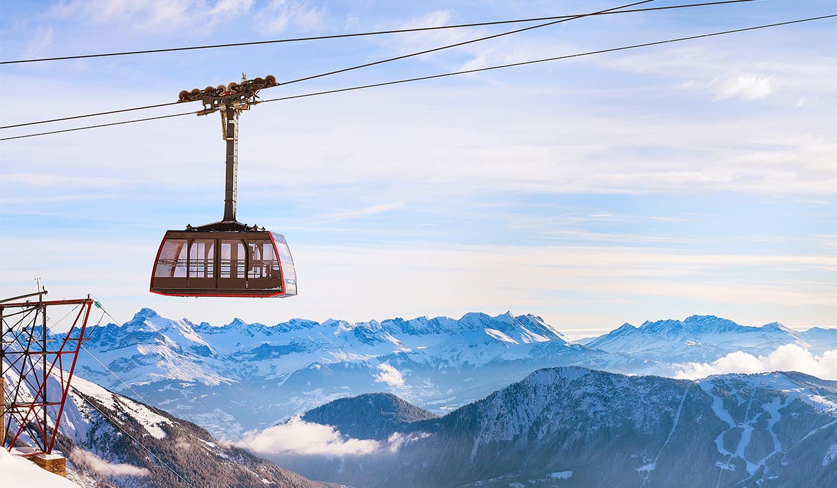 Aerial cable car suspended on a line above a snowy mountain range, with rugged peaks and clouds stretching across the horizon.