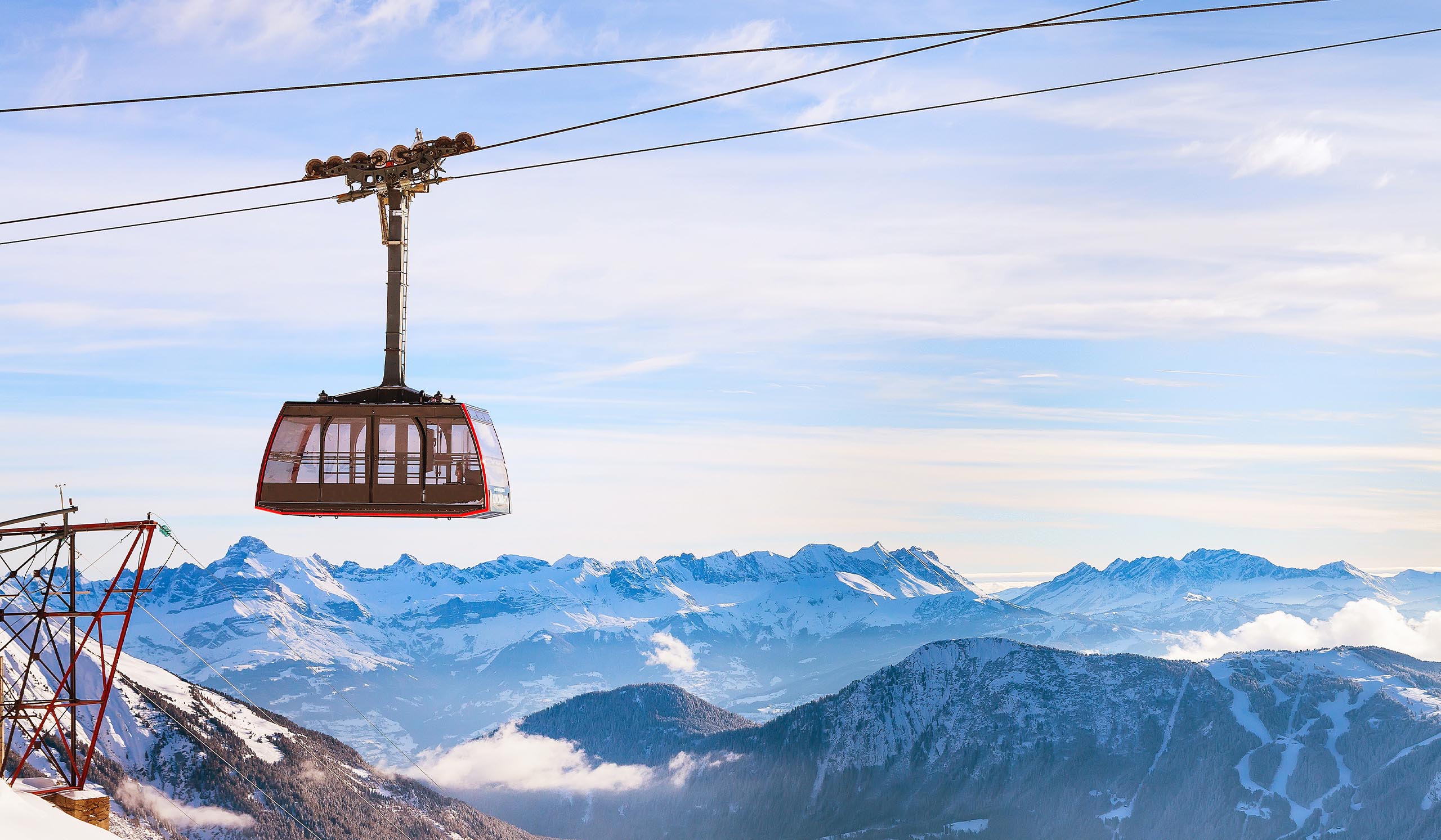 Aerial cable car suspended on a line above a snowy mountain range, with rugged peaks and clouds stretching across the horizon.