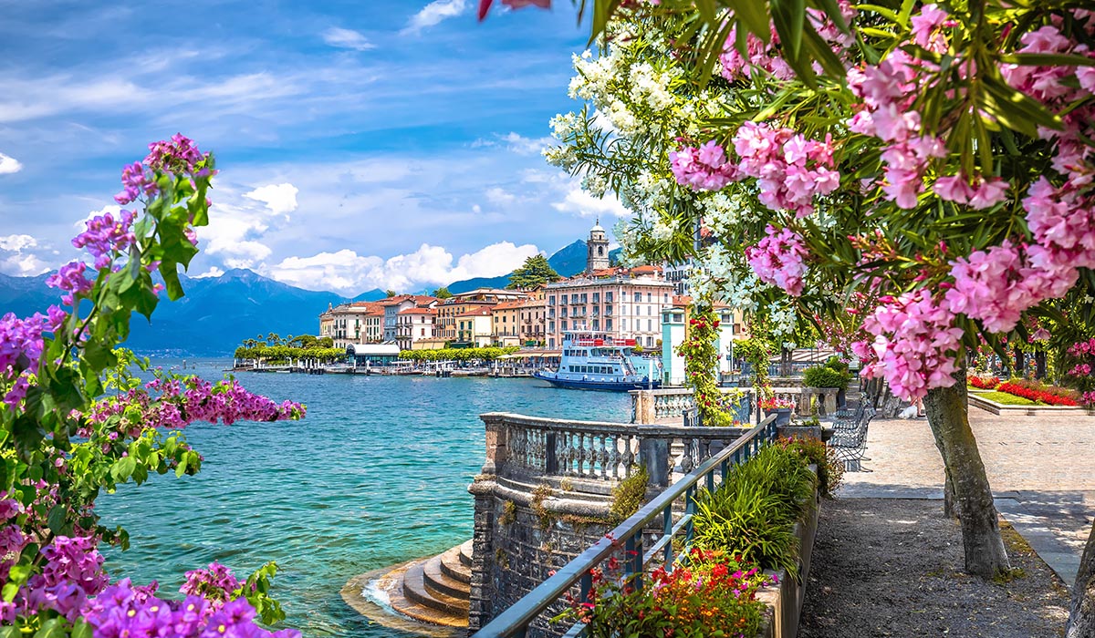 Charming lakeside town with pastel buildings and a waterfront promenade, framed by vibrant pink and white flowers, with mountains rising in the background under a bright blue sky.