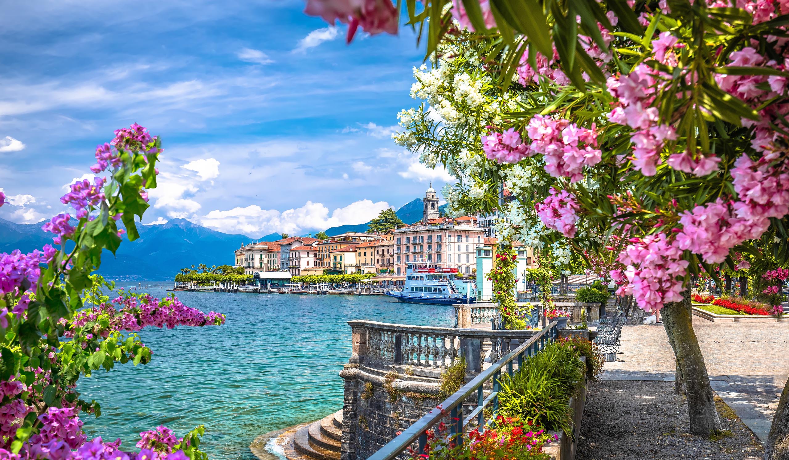 Charming lakeside town with pastel buildings and a waterfront promenade, framed by vibrant pink and white flowers, with mountains rising in the background under a bright blue sky.