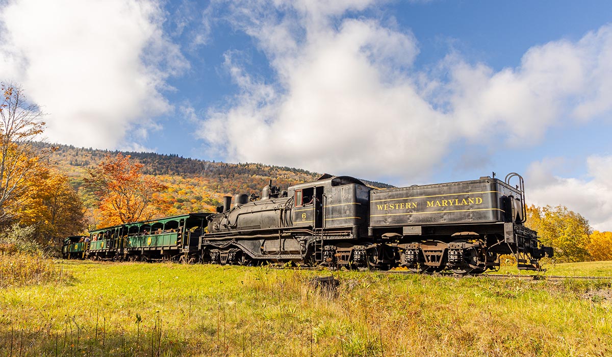 A black Western Maryland steam locomotive pulls green passenger cars through a grassy field, with colorful fall scnery covering the hills behind it under a partly cloudy blue sky.