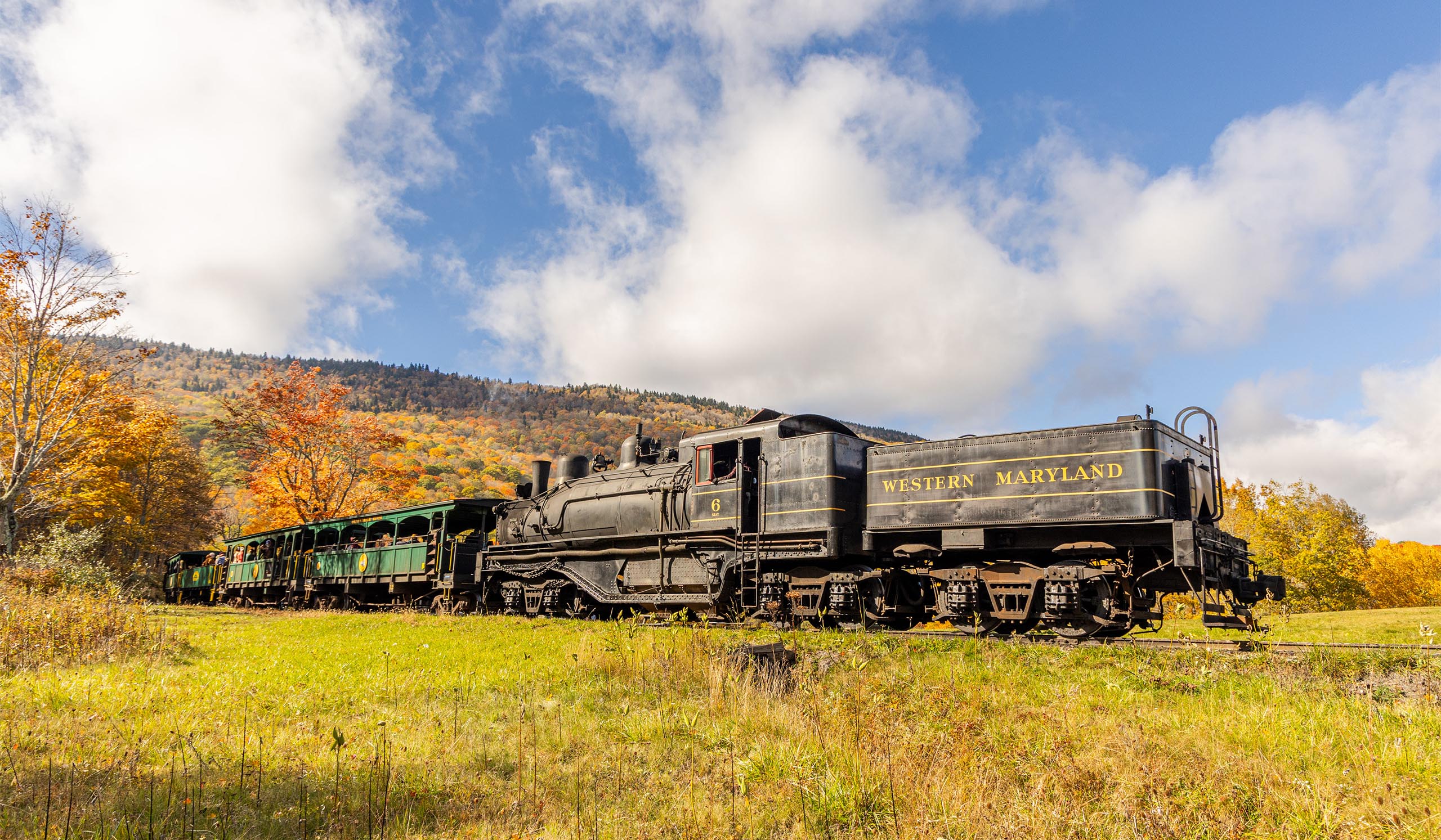 A black Western Maryland steam locomotive pulls green passenger cars through a grassy field, with colorful fall scnery covering the hills behind it under a partly cloudy blue sky.