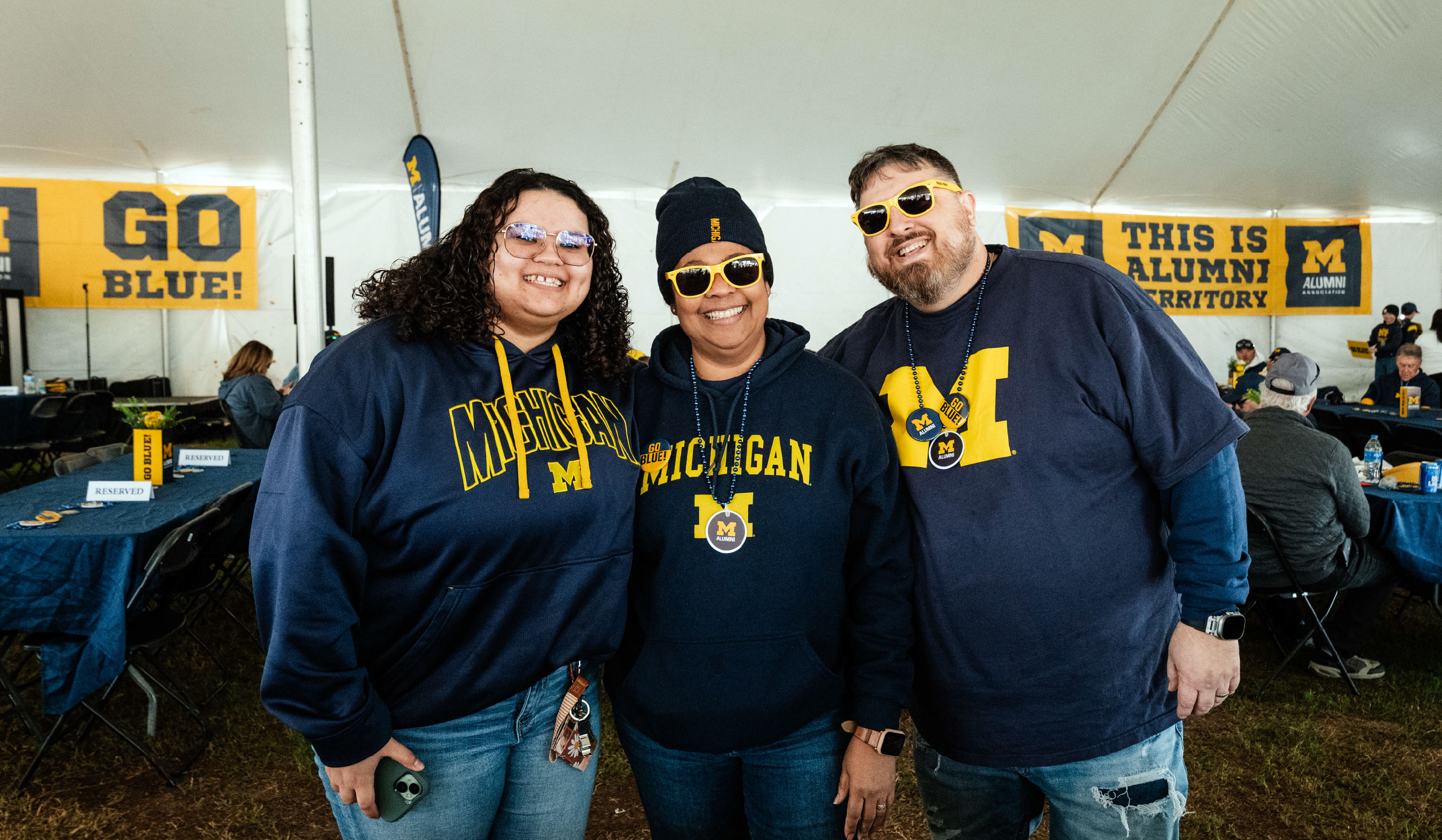 Three people in maize-and-blue Michigan apparel smile under a tent at an Alumni Territory tailgate.