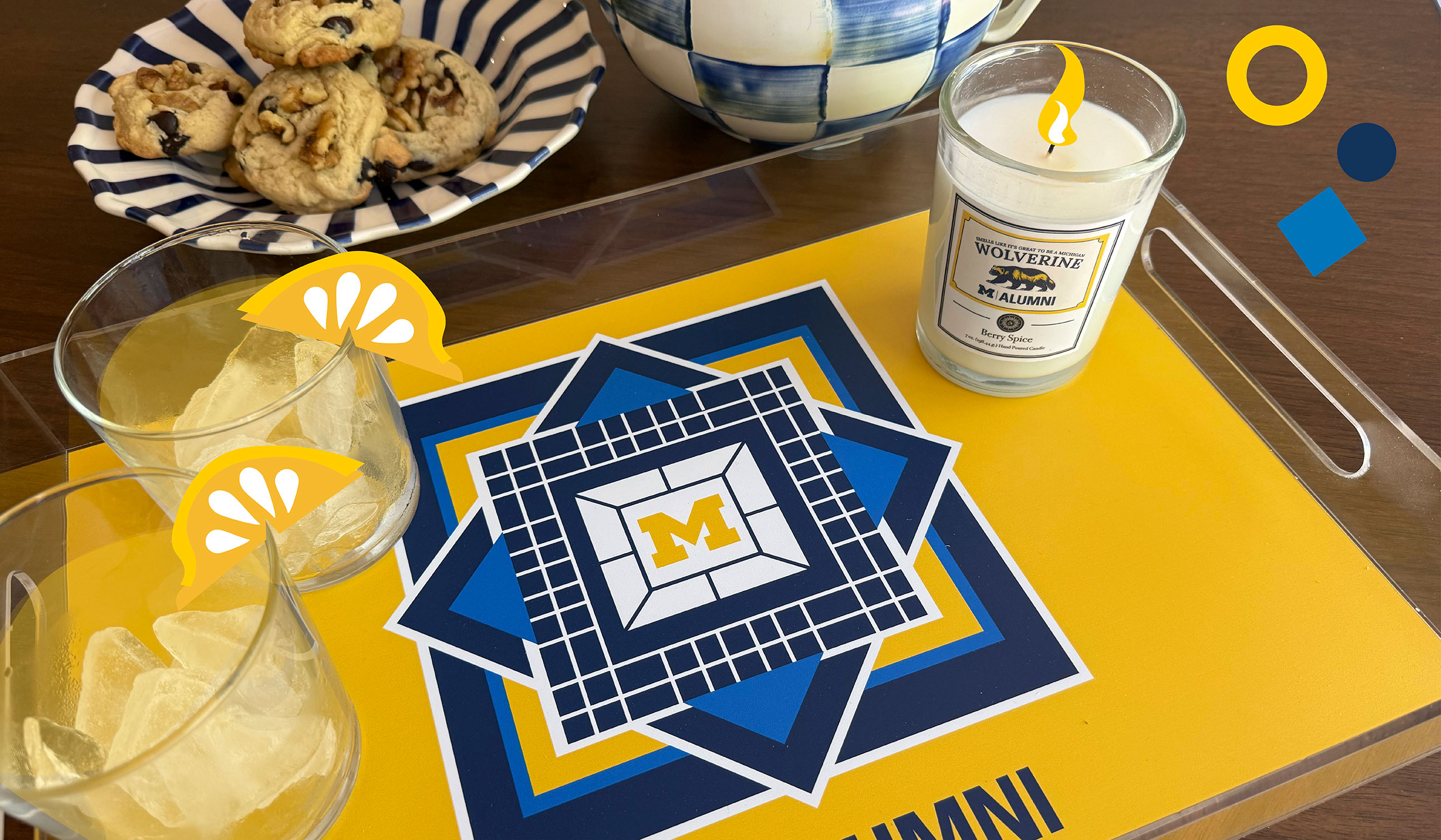 A maize-and-blue serving tray featuring a Block M in the center sits on a table. Two iced drinks are atop the tray, as is a Michigan-themed candle. A striped bowl with chocolate chip cookies is behind the tray.