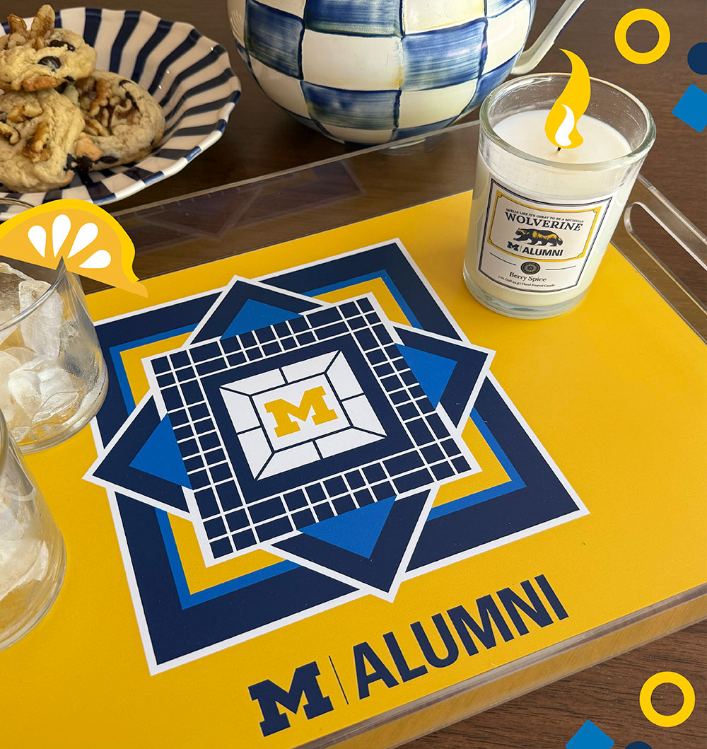 A maize-and-blue serving tray featuring a Block M in the center and M Alumni at the bottom sits on a table. Two iced drinks are atop the tray, as is a Michigan-themed candle. A striped bowl with chocolate chip cookies is behind the tray.