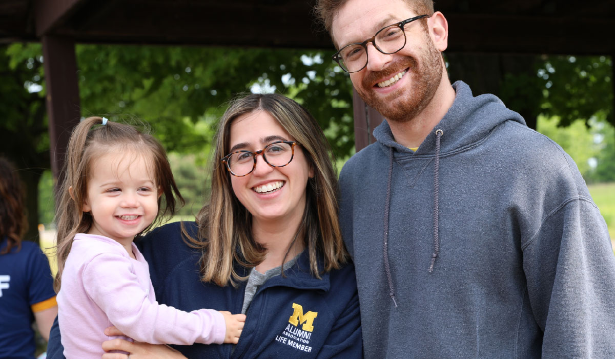 A person in an Alumni Association of the University of Michigan Life Member fleece smiles next to her husband while holding her child.