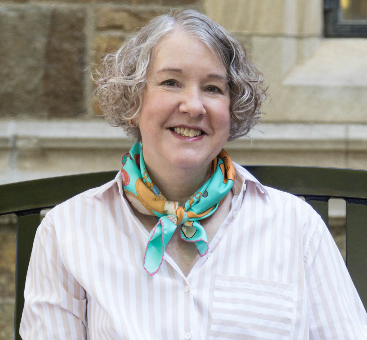 Laura Lamps has short, curly gray hair as she sits on a dark bench, dressed in a pale pink and white striped shirt with an aqua and yellow scarf tied at the neck; a stone wall serves as the background.