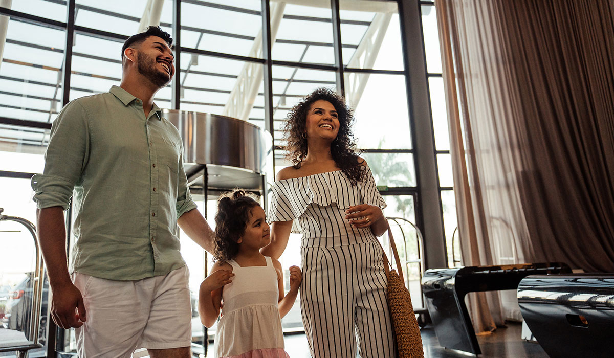 A man, woman, and young girl walk side by side in a bright hotel lobby with large windows and modern decor. The man is wearing a light shirt and shorts, the woman is dressed in a striped outfit and carrying a woven bag, and the girl is in a white dress.