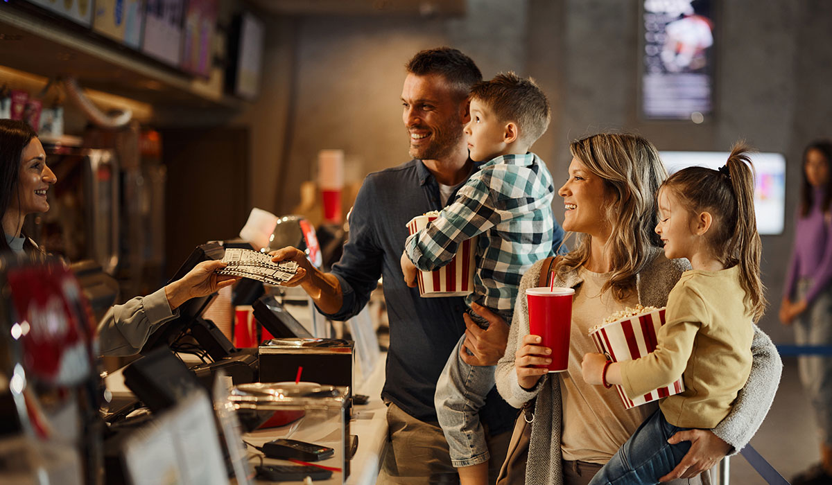 Two adults and two children stand at a movie theater concession counter. The adults are holding the kids, who each have popcorn, and there are drinks in hand as they interact with the cashier.