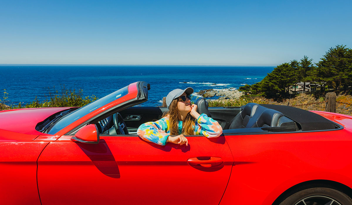 A person wearing a tie-dye sweatshirt and gray baseball cap is seated in a bright red convertible, parked near a scenic coastline with blue water and trees. It’s sunny with clear skies.