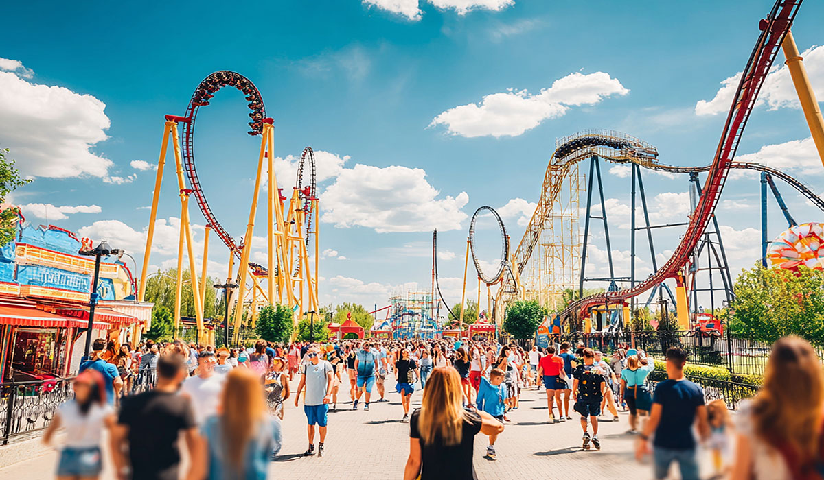 Groups of people stroll along a wide pathway in a bustling amusement park, surrounded by tall roller coasters and colorful buildings, all beneath vibrant clouds and sunshine.