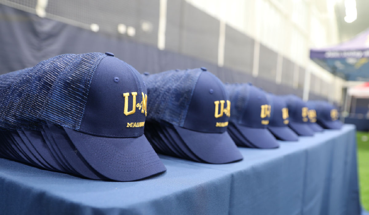 A row of navy hats stacked on a table. The hats feature U of M and an M Alumni logo in maize on the front of them.