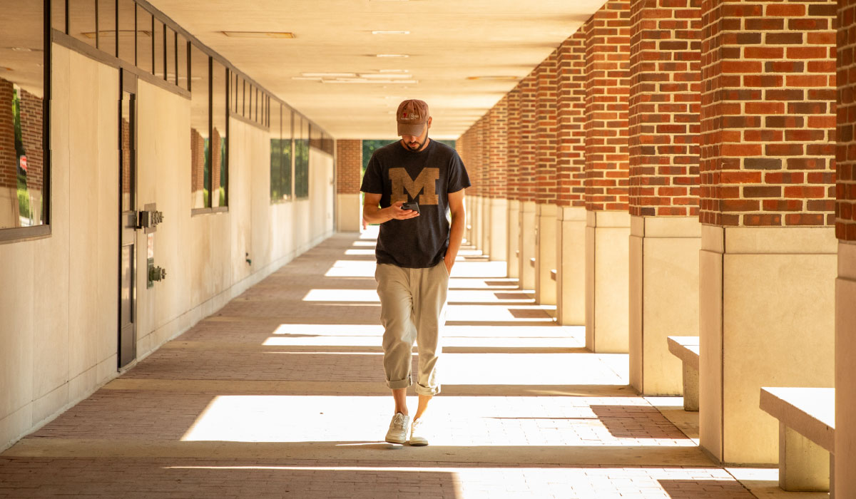 A person in a Block M shirt looks down at their phone as they walk through a brick-lined walkway toward the camera.