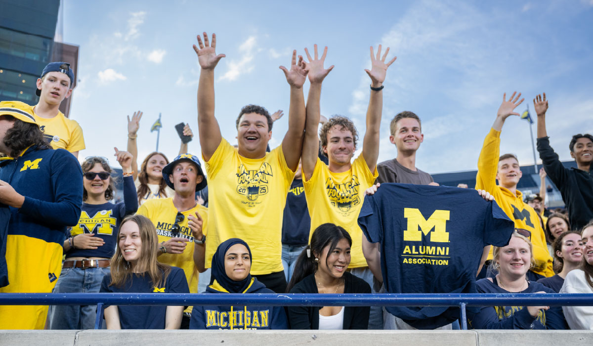 A crowd of University of Michigan students in maize and blue at Michigan Stadium, with several of them raising their arms in the air. One student holds up an Alumni Association of the University of Michigan shirt.