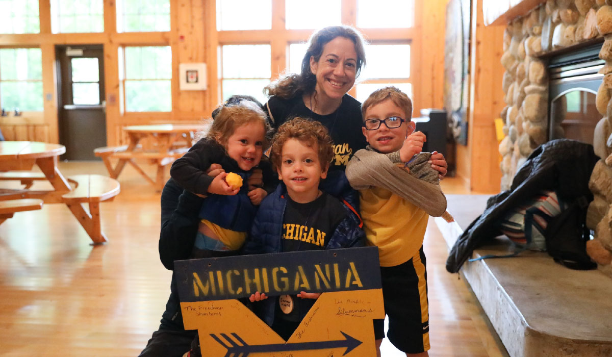 An adult and three kids in maize-and-blue Michigan apparel smile behind a Camp Michigania sign.