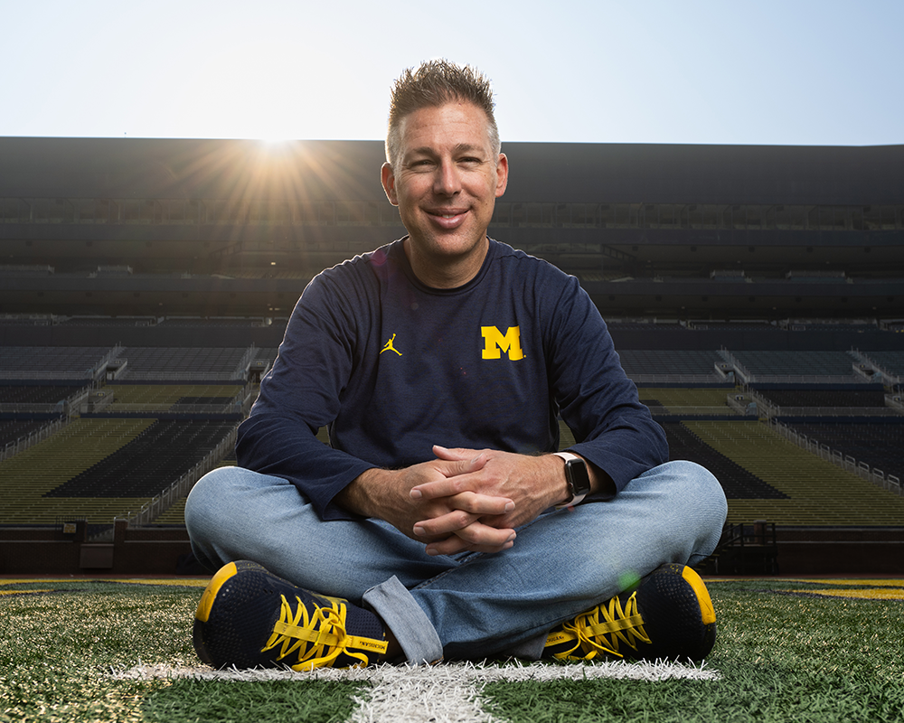 John Pasquale sitting on the field at Michigan Stadium wearing a blue shirt with a maize block M.