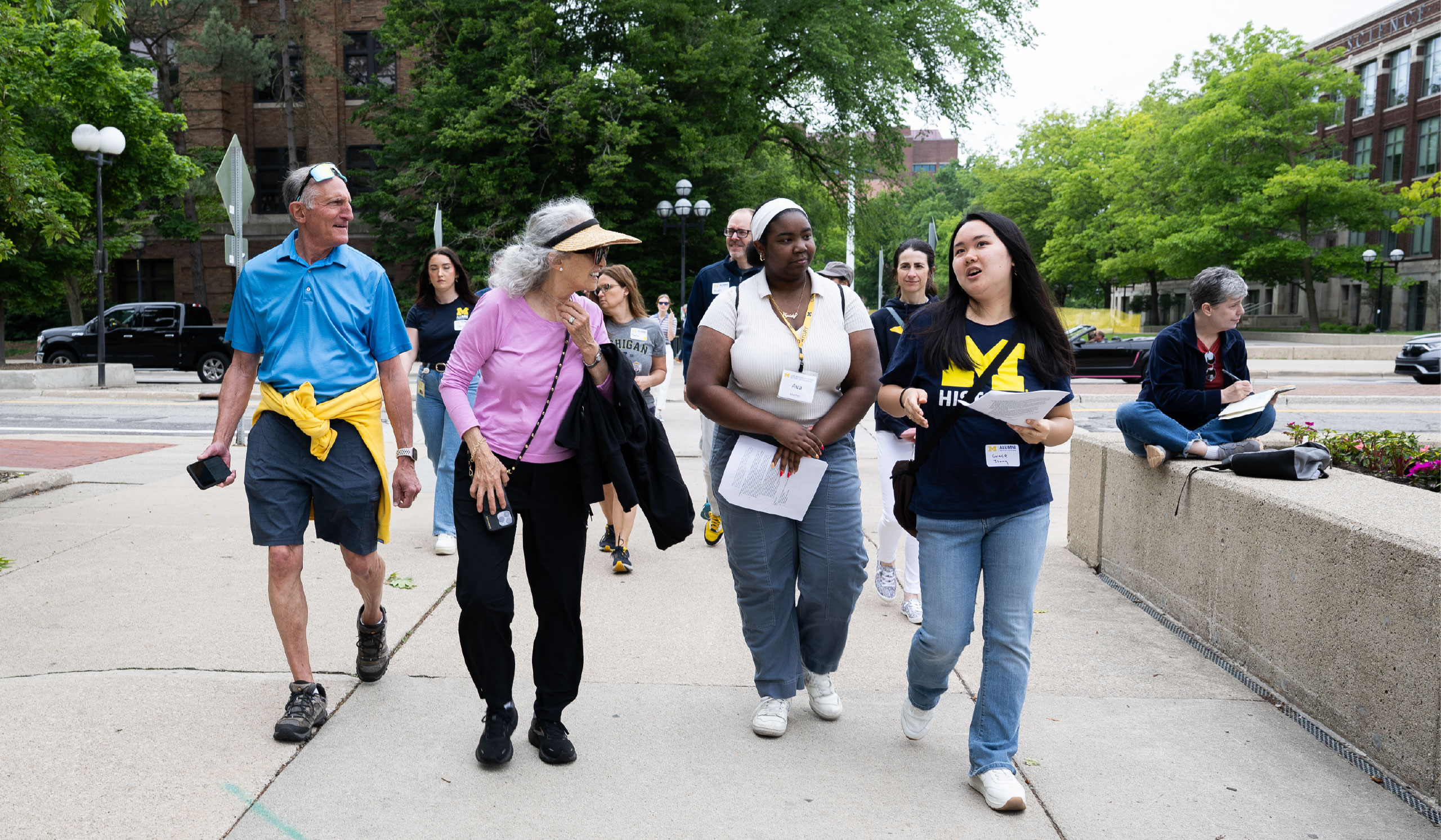 Two University of Michigan students lead a group of alumni on a tour across the Ann Arbor campus. Lush green trees cover the background.