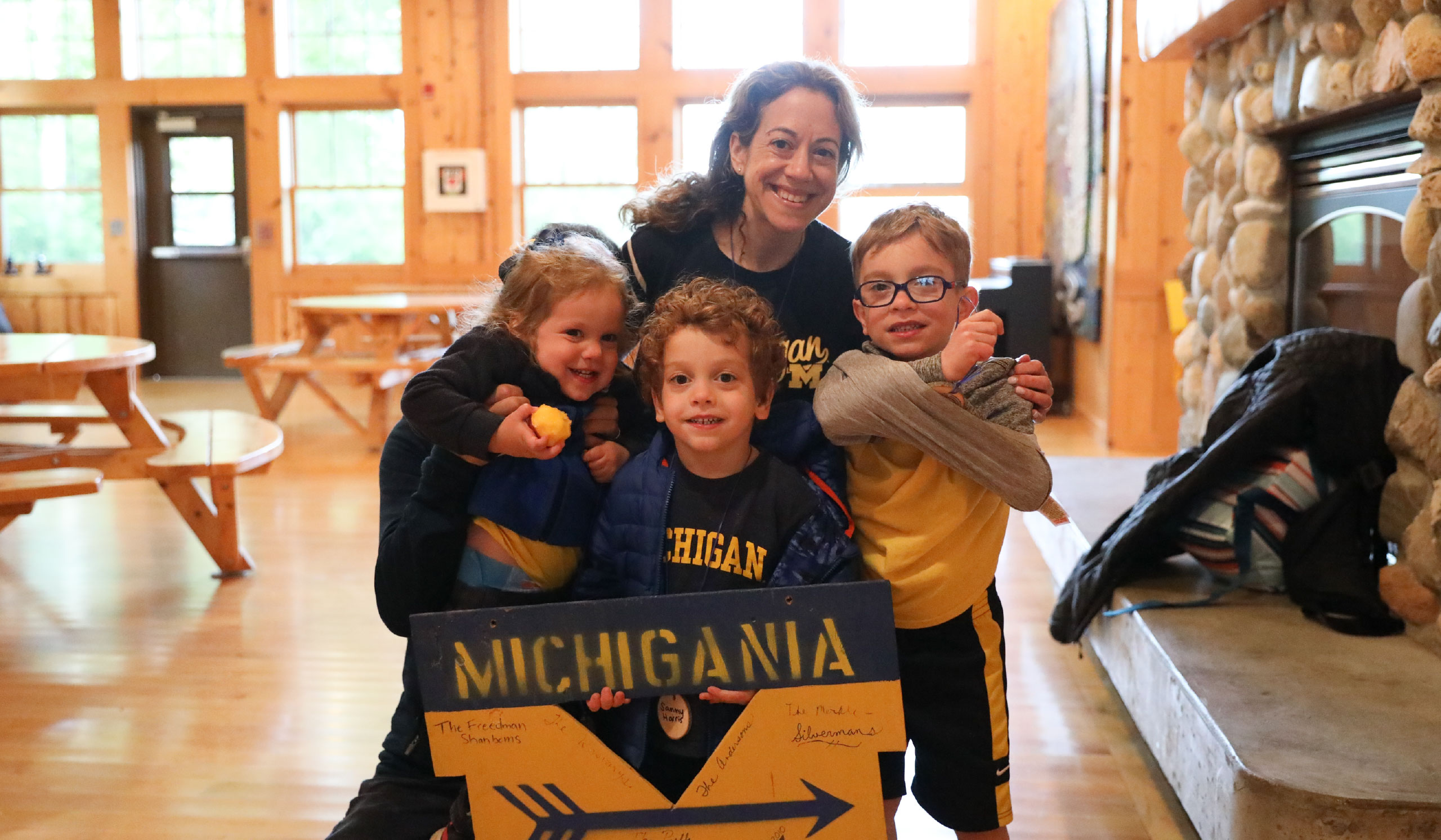 An adult and three kids in maize-and-blue Michigan apparel smile behind a Camp Michigania sign.