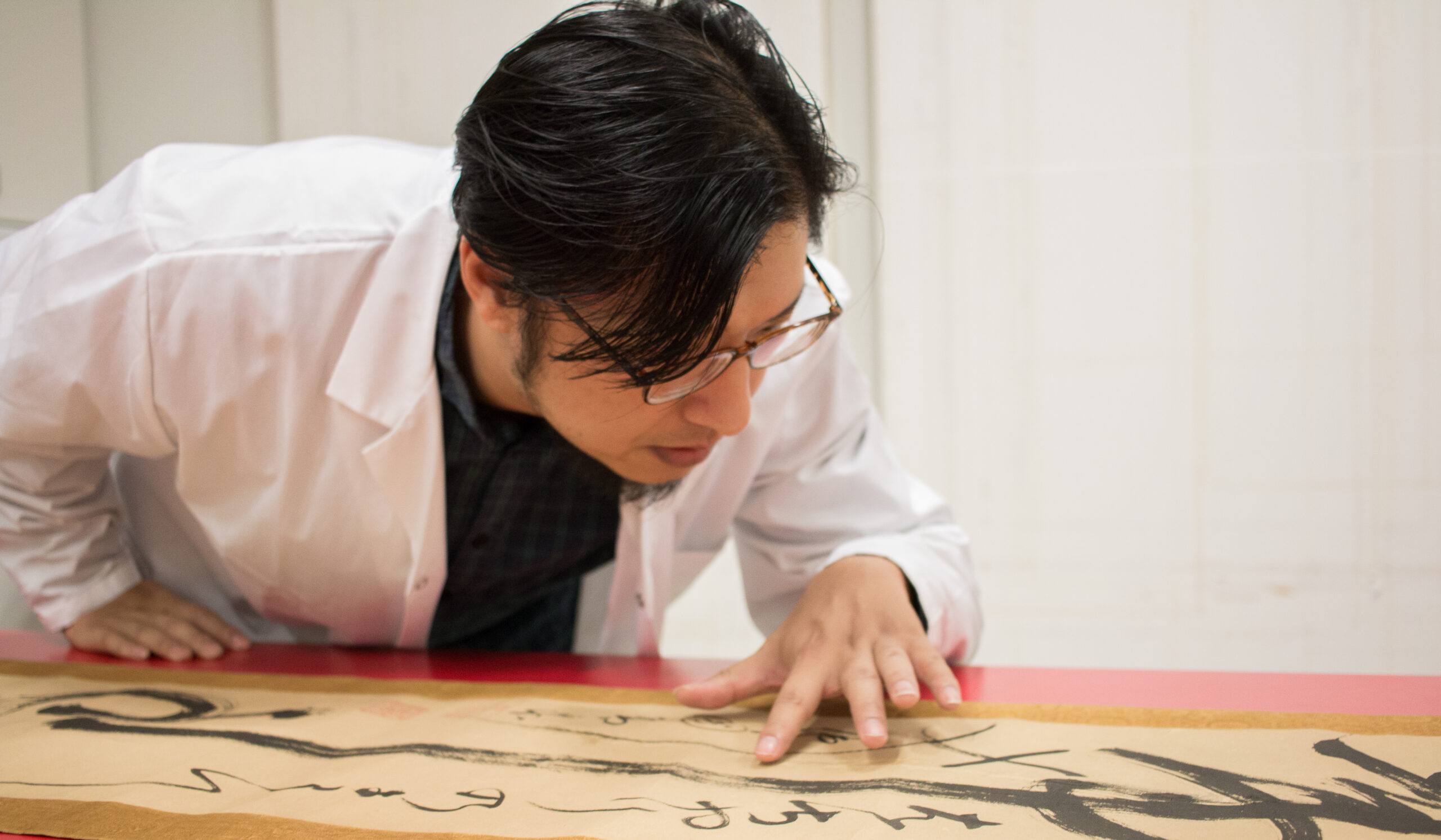 A person in a white lab coat and glasses studies a long scroll with black calligraphy, hand and face near the artwork on a red table.
